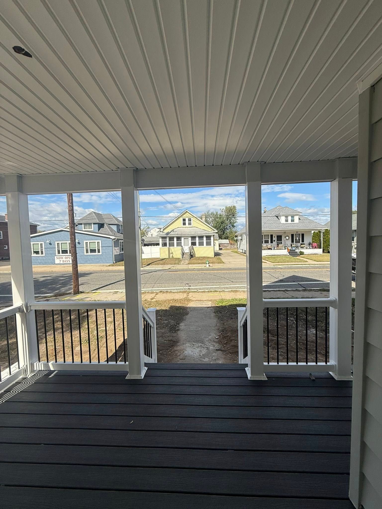 View from a porch with white pillars, overlooking a street with houses and blue sky.