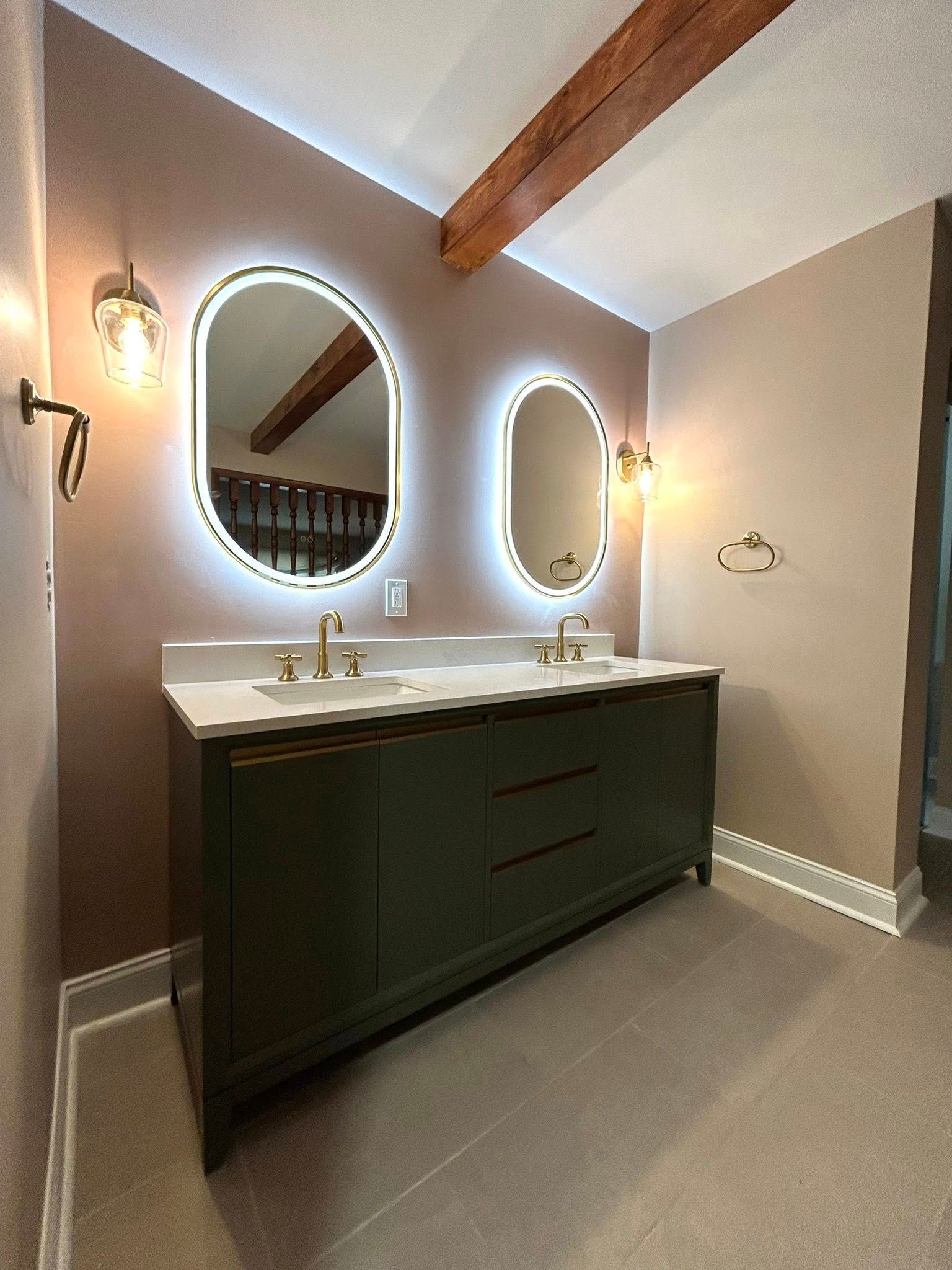 Green double vanity with oval mirrors, sconces, and wood beam in a bathroom.