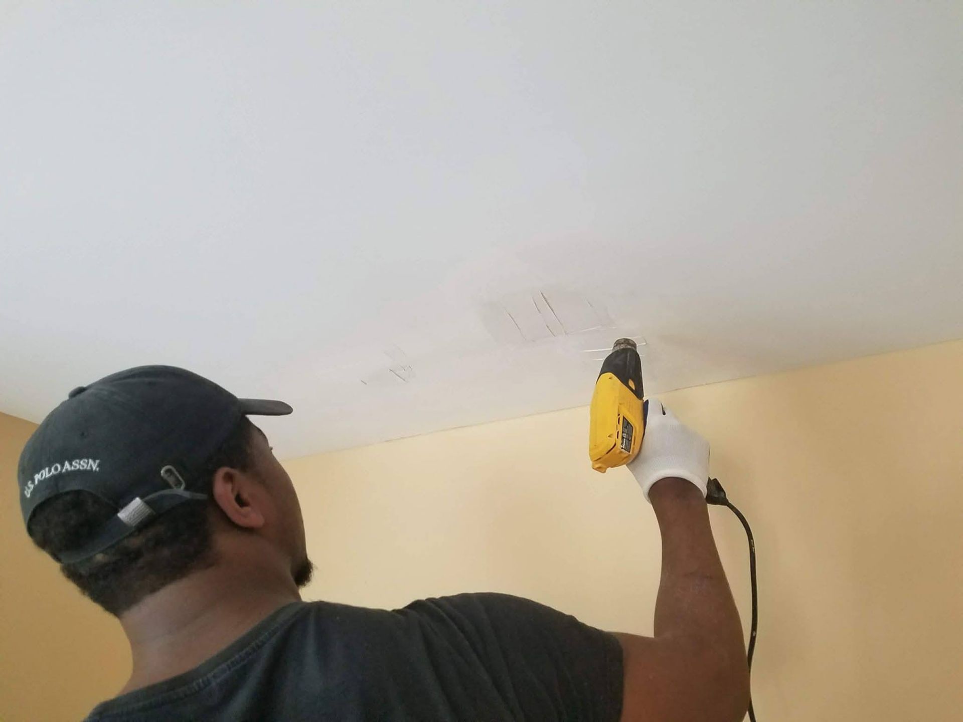A person wearing a hat and a glove uses a handheld yellow rotary tool to cut into a white ceiling.