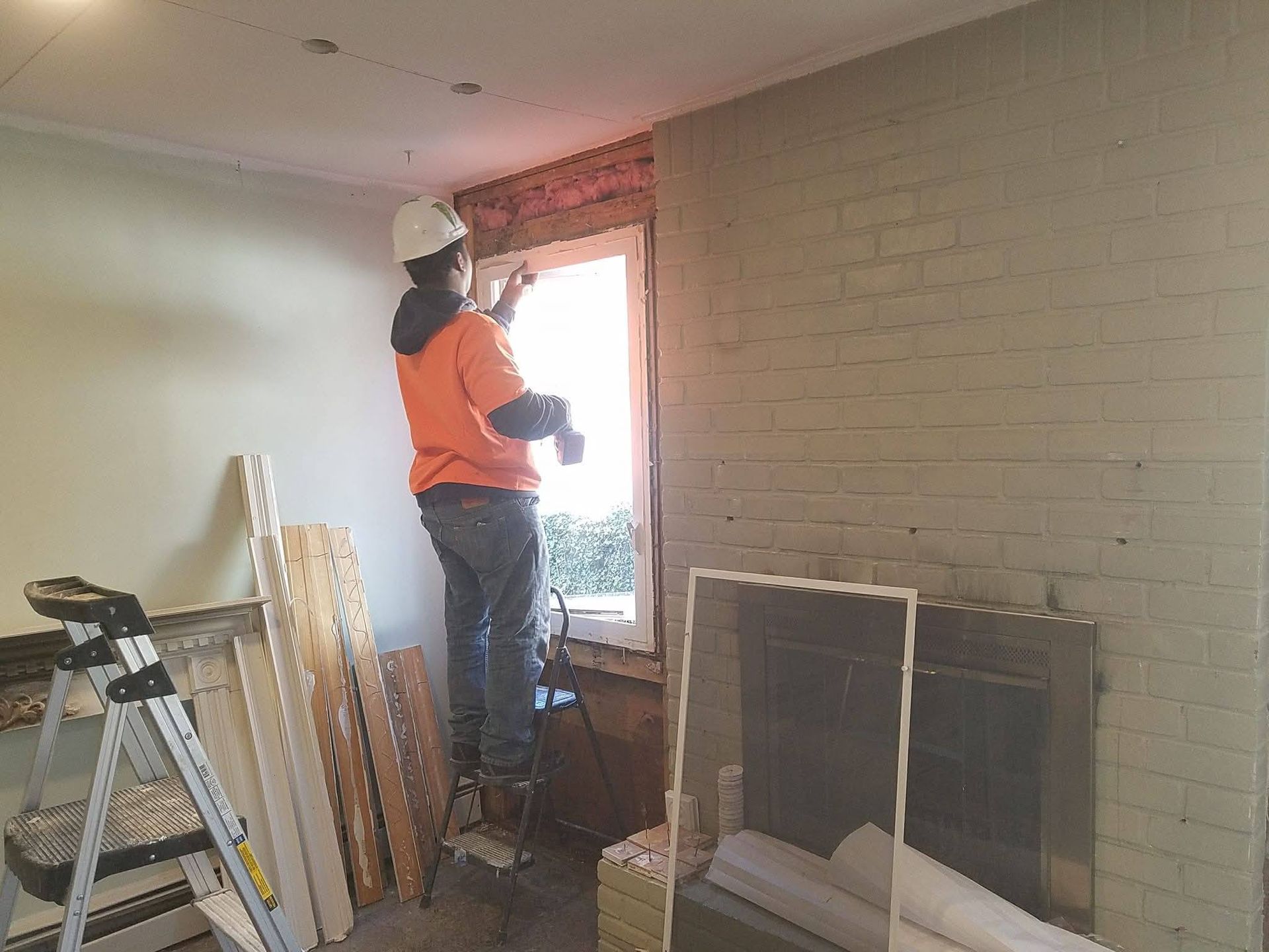 A construction worker in a high-visibility orange jacket stands on a ladder, installing or repairing a window frame.