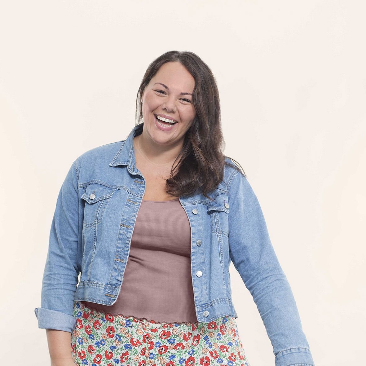 A woman wearing a denim jacket and a floral skirt is smiling.