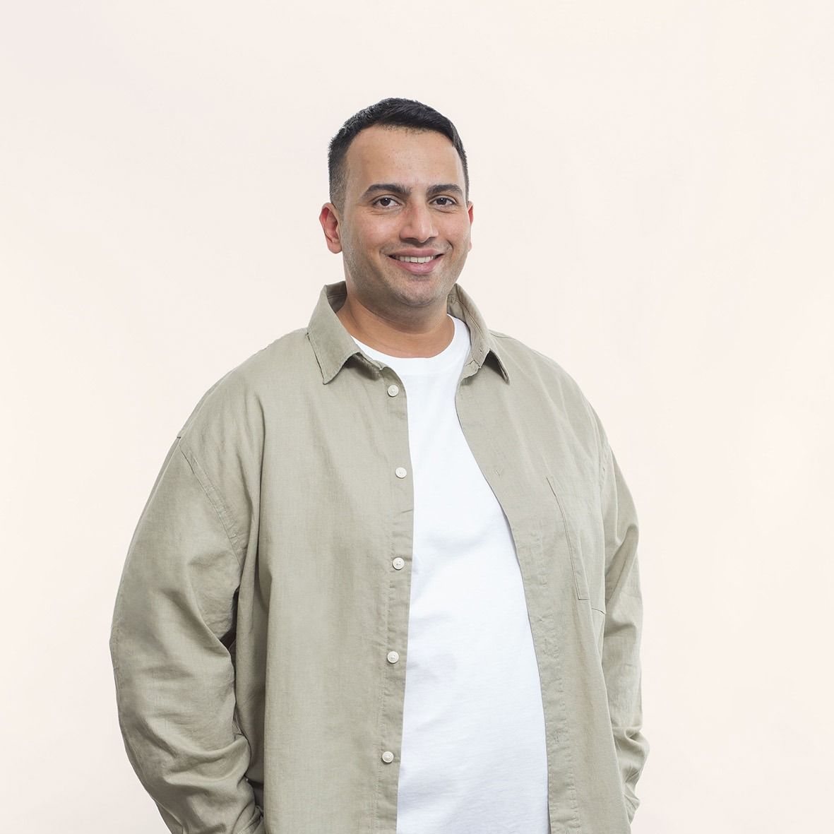 A man wearing a tan shirt and a white shirt is standing with his hands in his pockets.