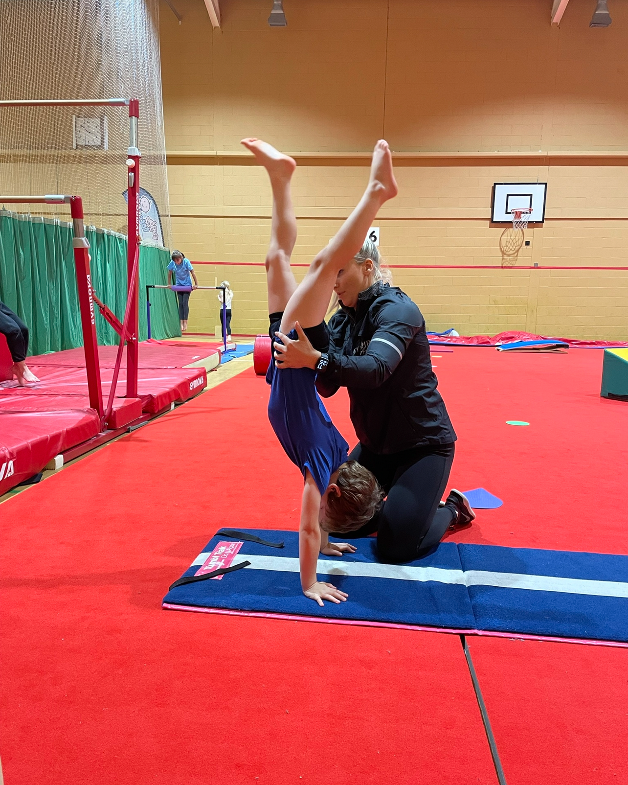 A man is helping a young boy do a handstand on a mat in a gym.
