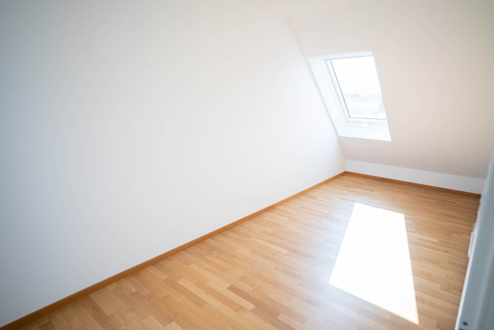 Leerer Raum mit Holzboden, weißen Wänden und einer Dachschräge mit Oberlicht.