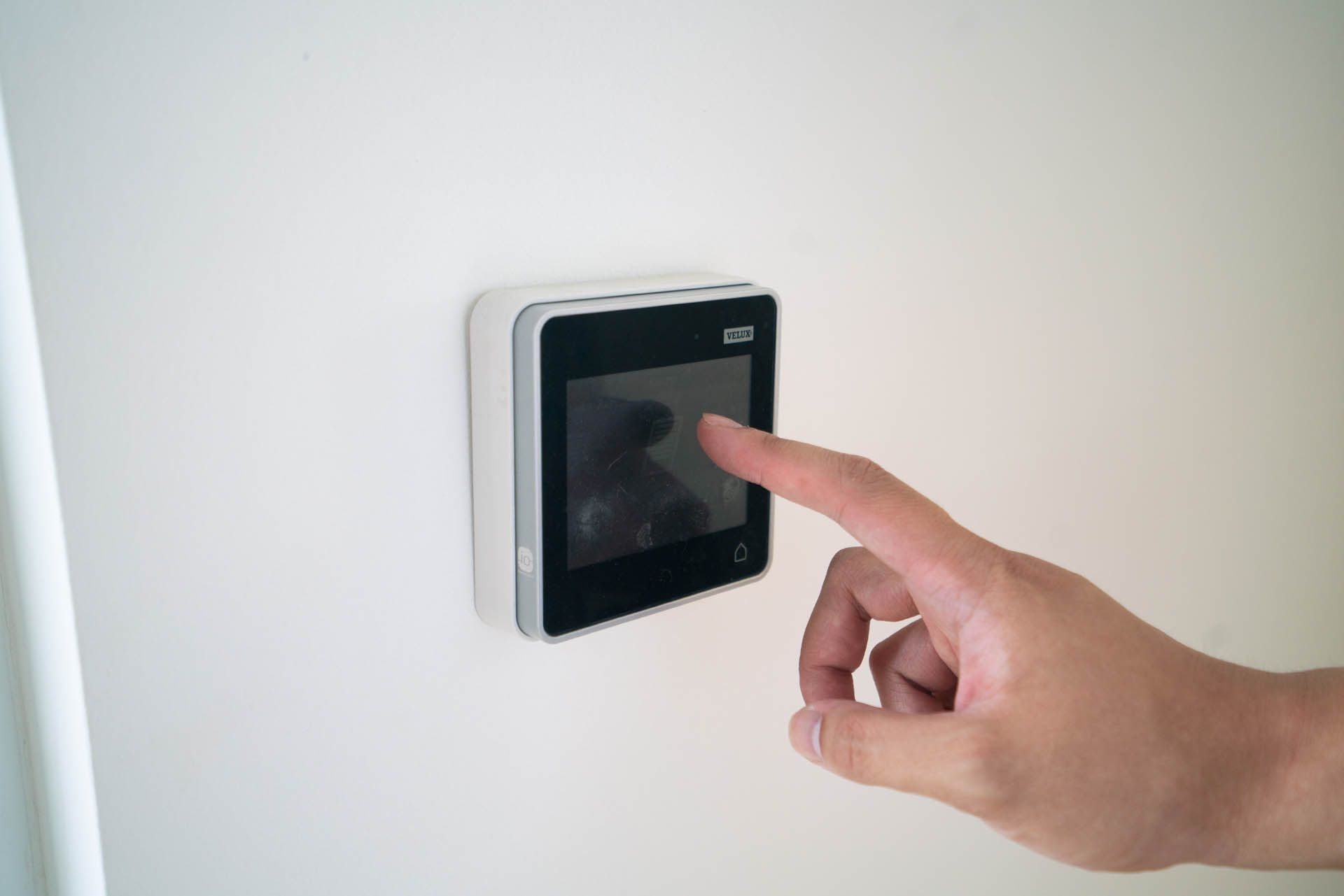 Eine Hand berührt ein quadratisches Gerät mit schwarzem Bildschirm an einer weißen Wand.