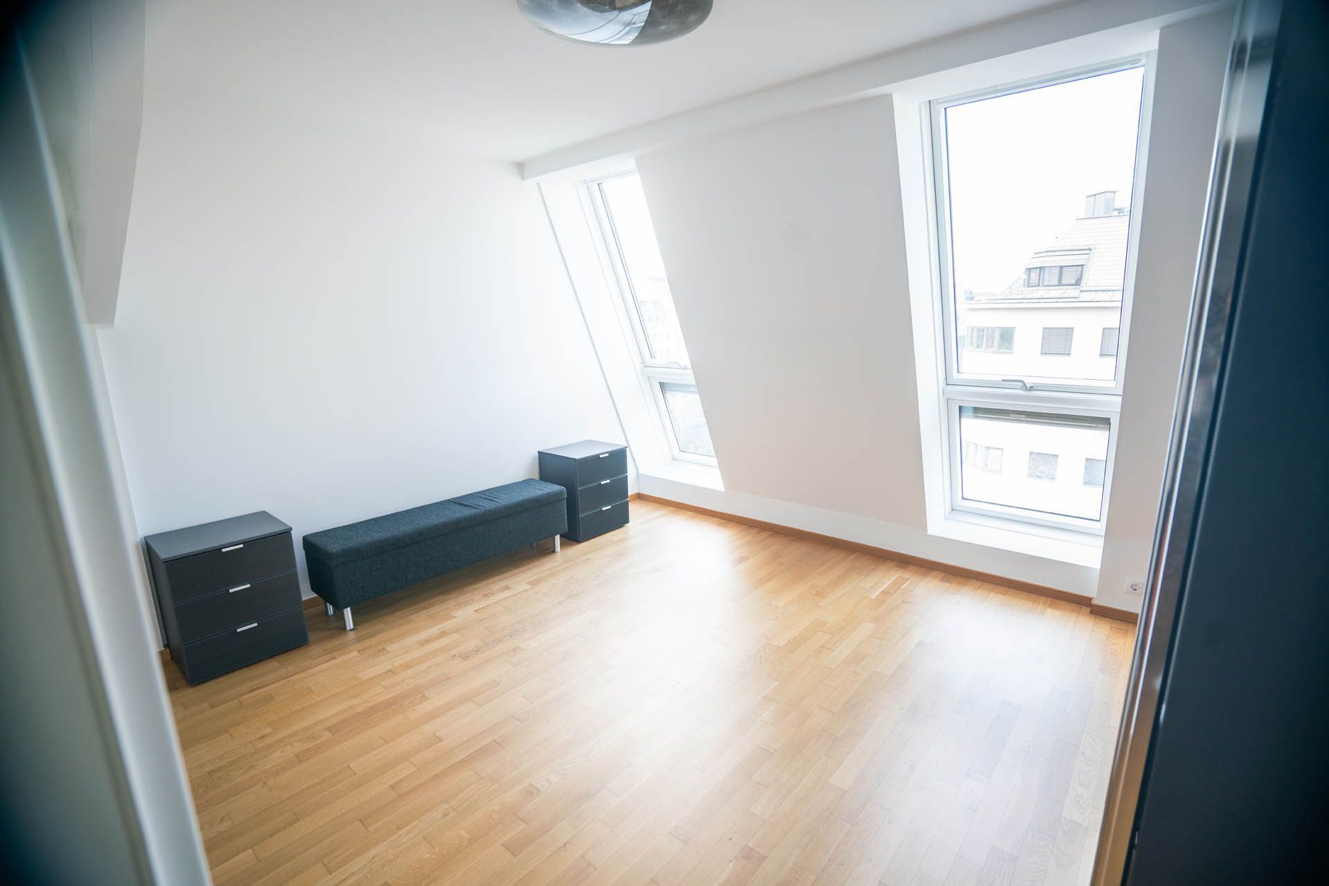 Leeres Schlafzimmer mit Holzboden, weißen Wänden, schwarzen Möbeln und großen Fenstern.