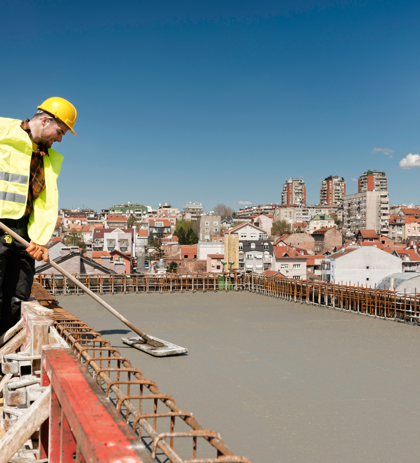 Construction worker smoothing concrete on a rooftop, city in background, bright sky.