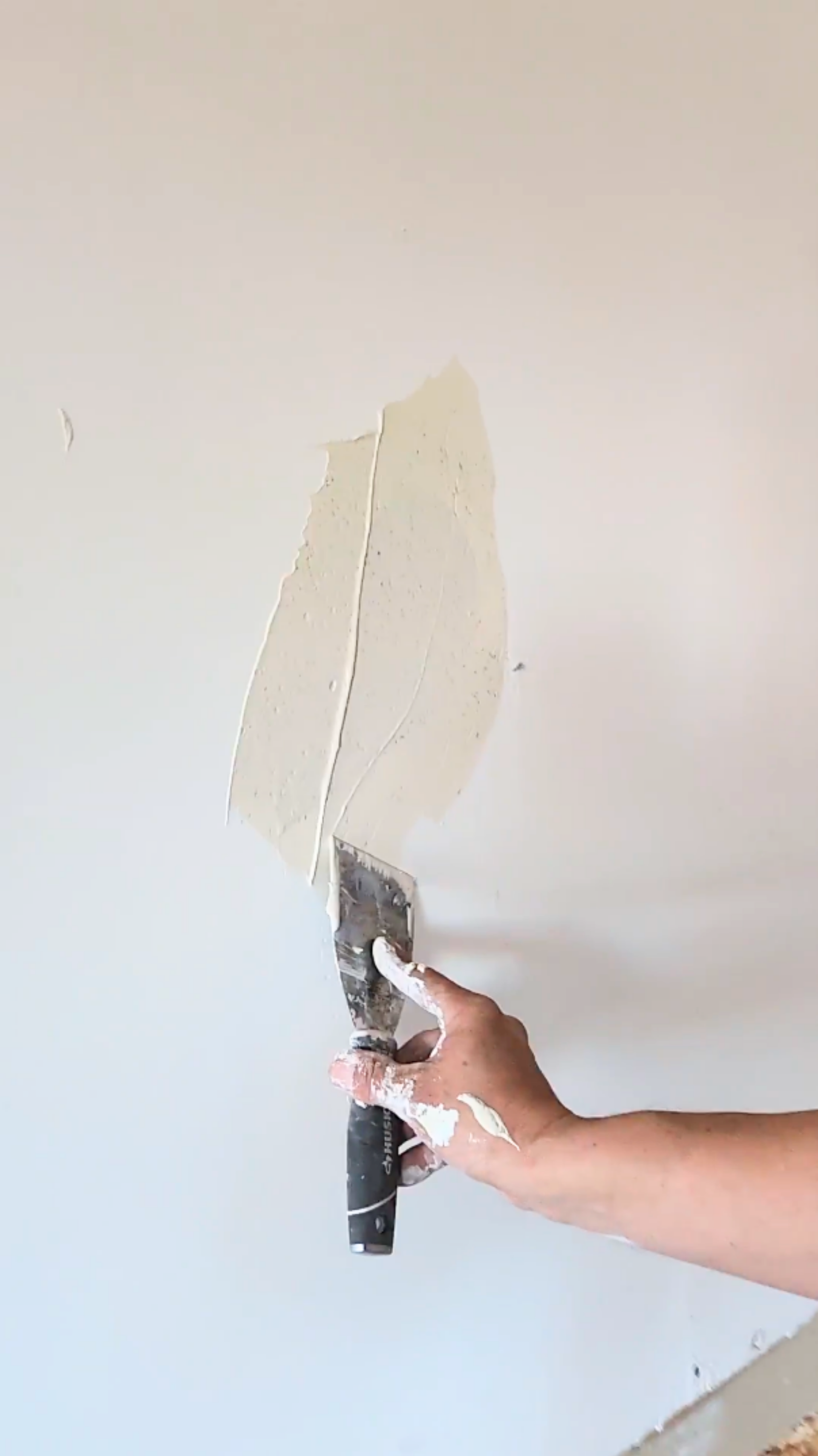 Hand using a putty knife to apply white spackle to a white wall.