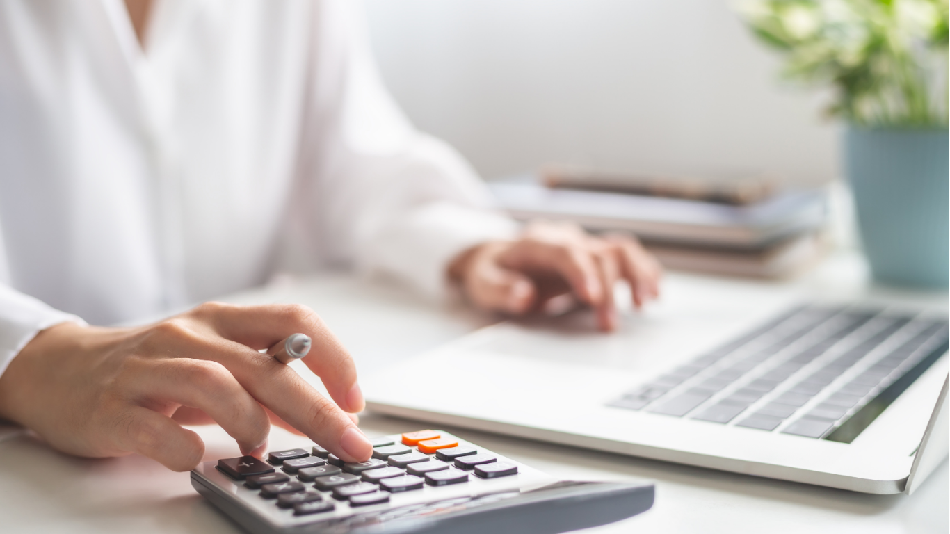 Woman's hands using a calculator at a desk with a laptop and notebook.