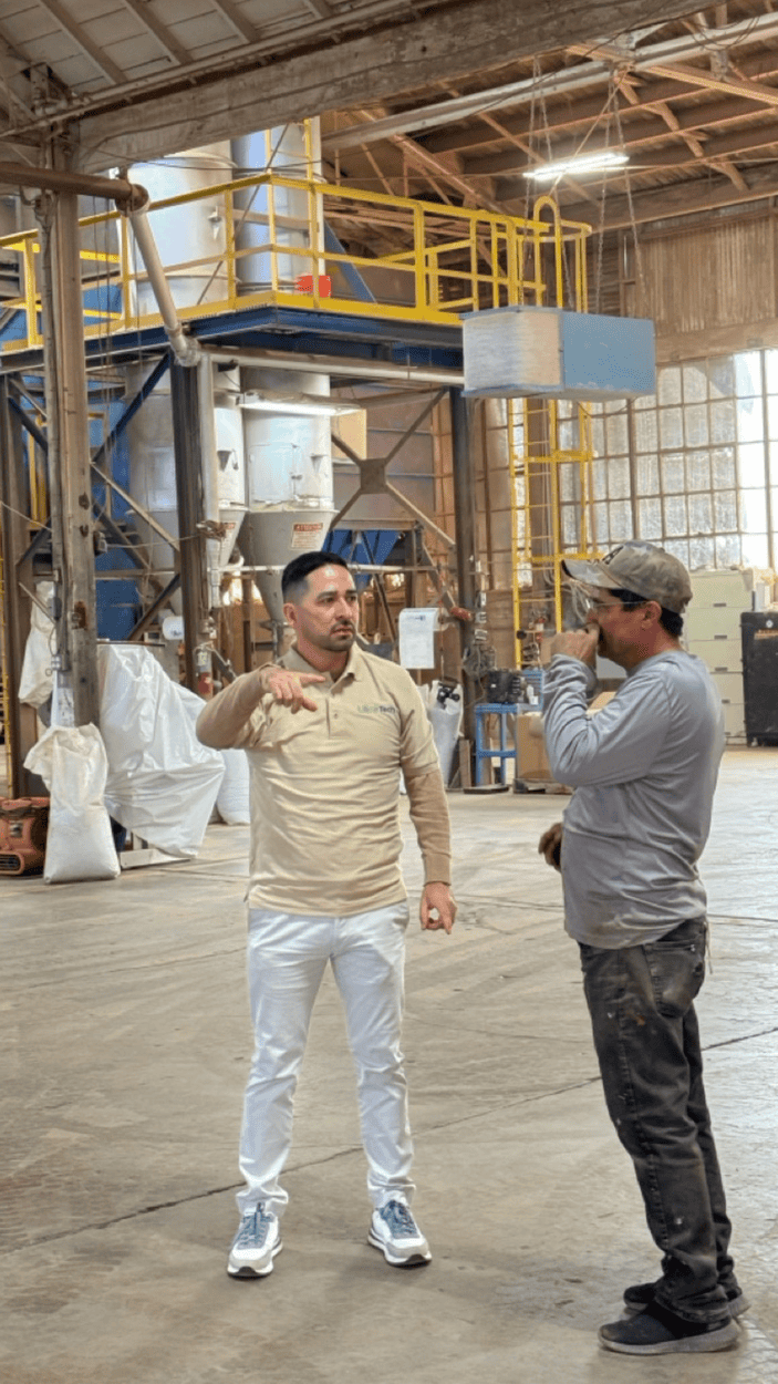 Two men in a warehouse; one gestures, the other listens. Industrial machinery is in the background.