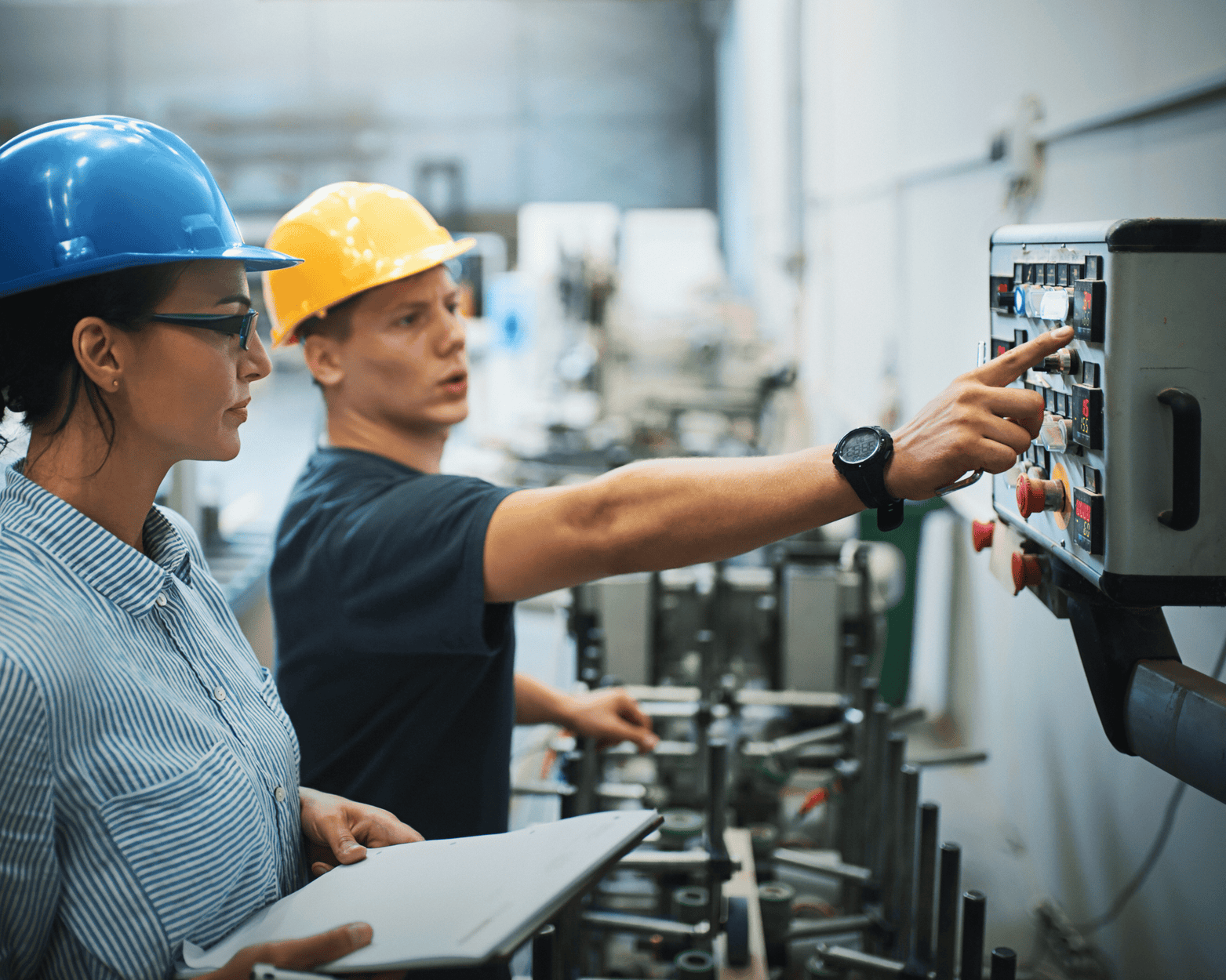 Woman and man in hard hats operating machinery in a factory, one pointing at a control panel.