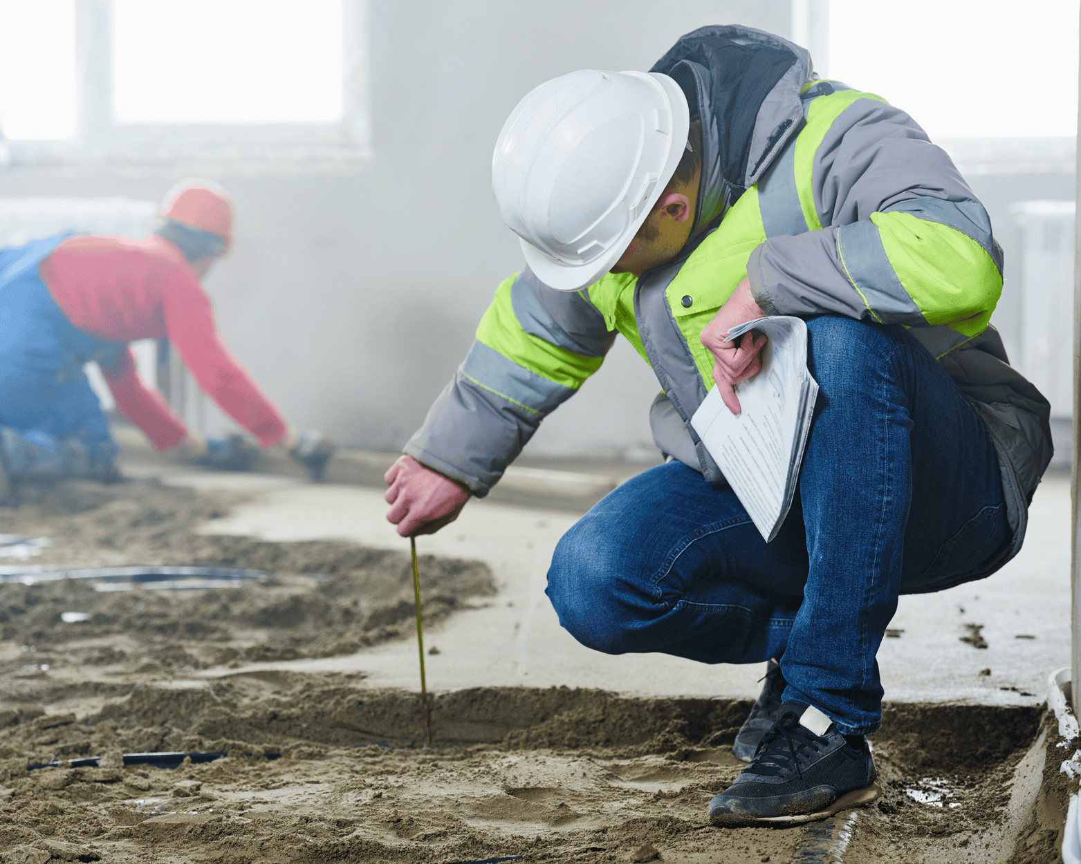 Construction worker inspecting floor, wearing white helmet and reflective vest, with another worker in the background.