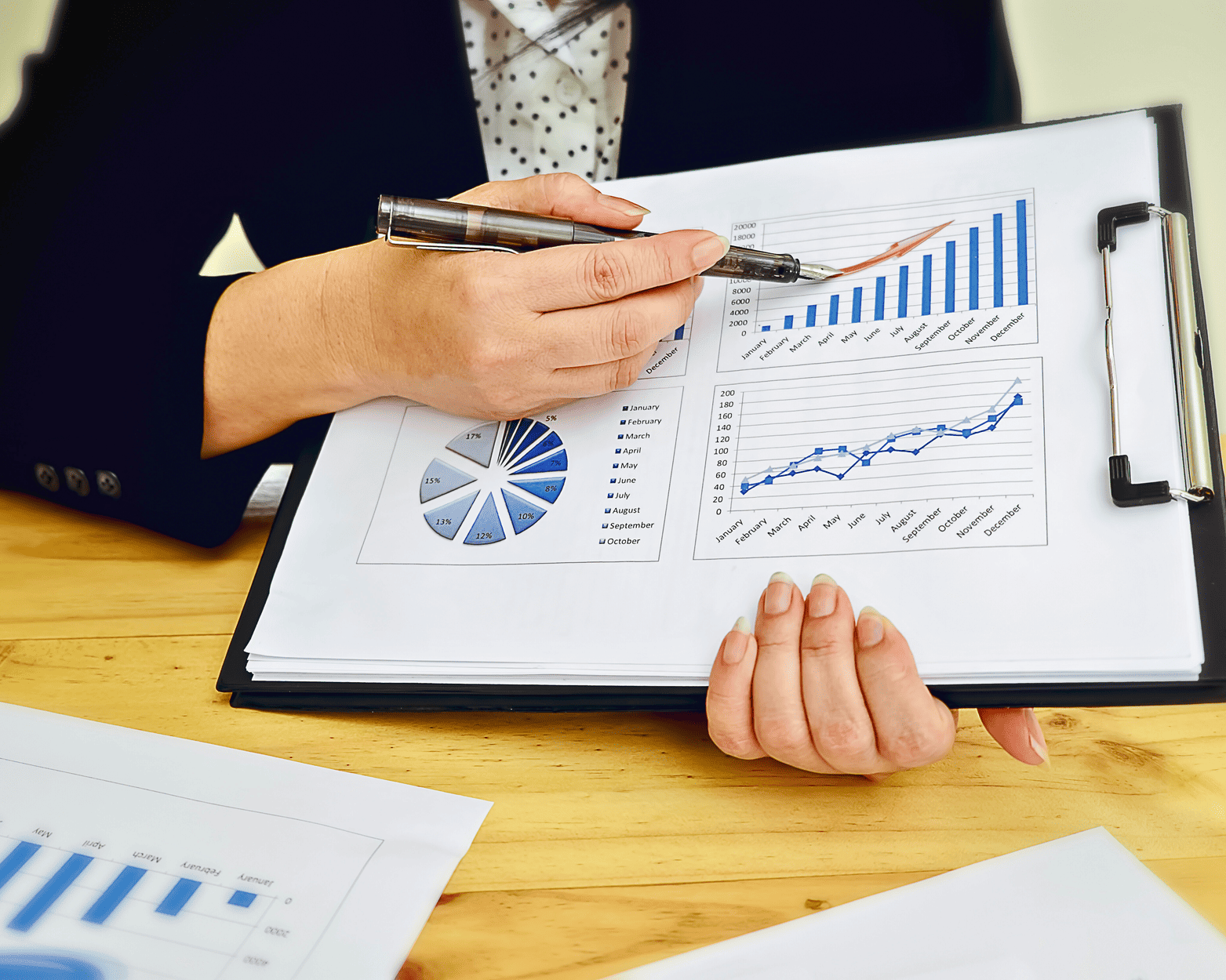 Person in suit points to financial charts on a clipboard; wooden desk.
