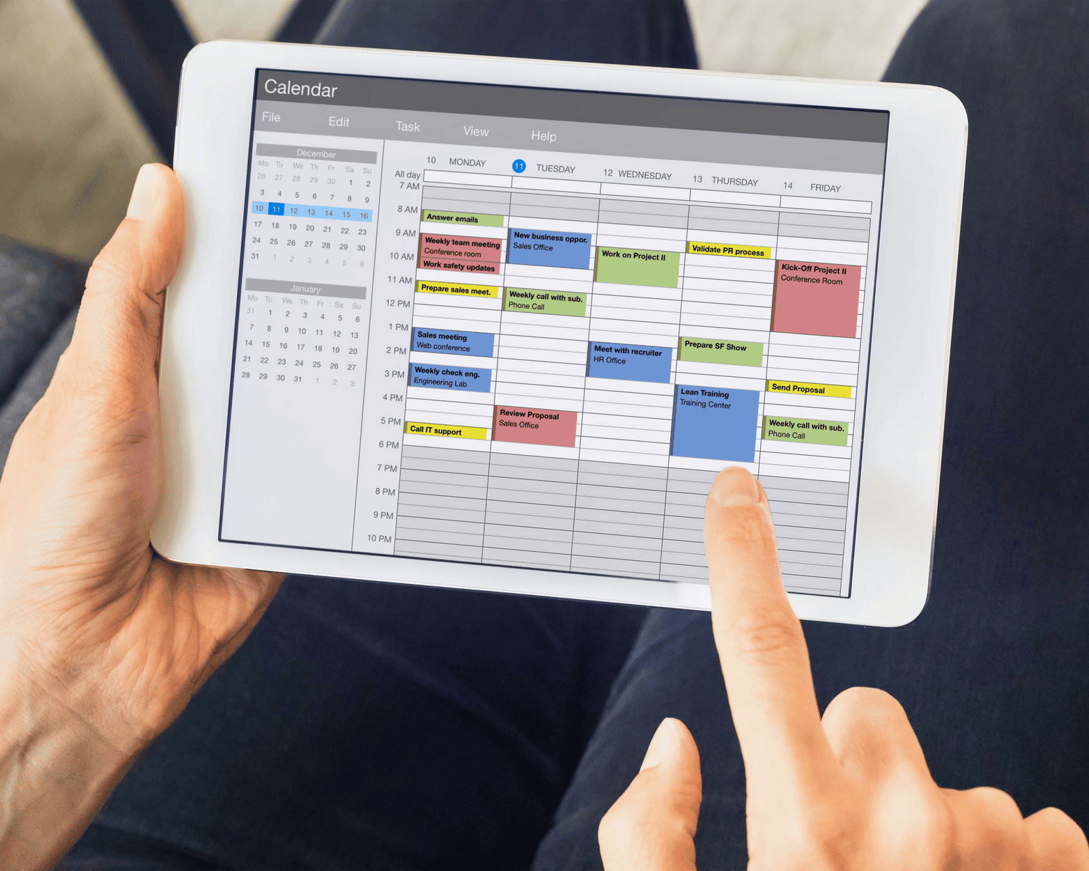 Person holding a tablet displaying a color-coded calendar, using finger to interact with the screen.