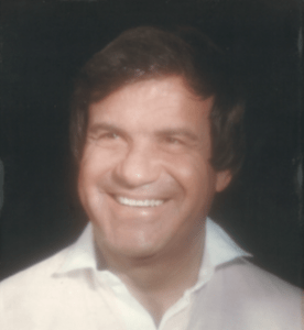 Smiling man with dark hair, wearing a white collared shirt, against a dark background.