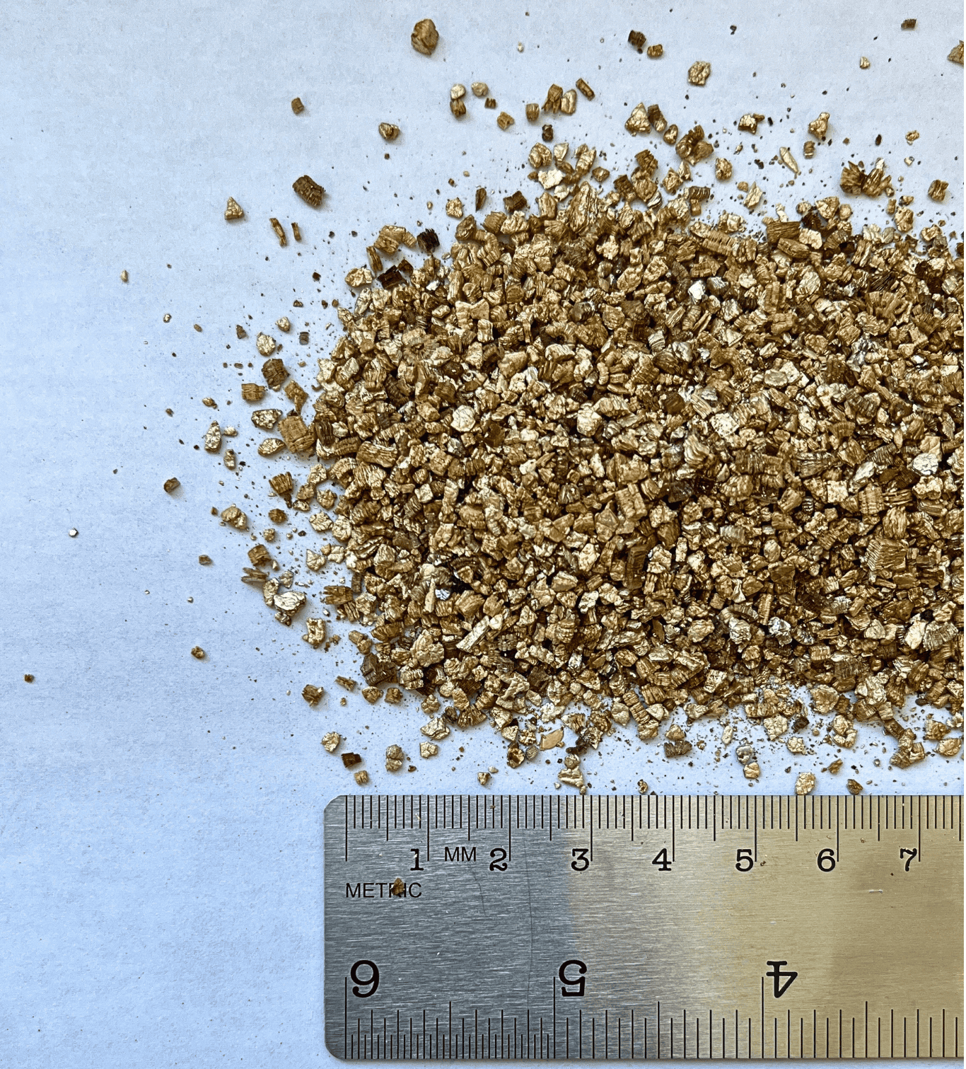 Brown vermiculite aggregate pile next to a ruler on a white surface.