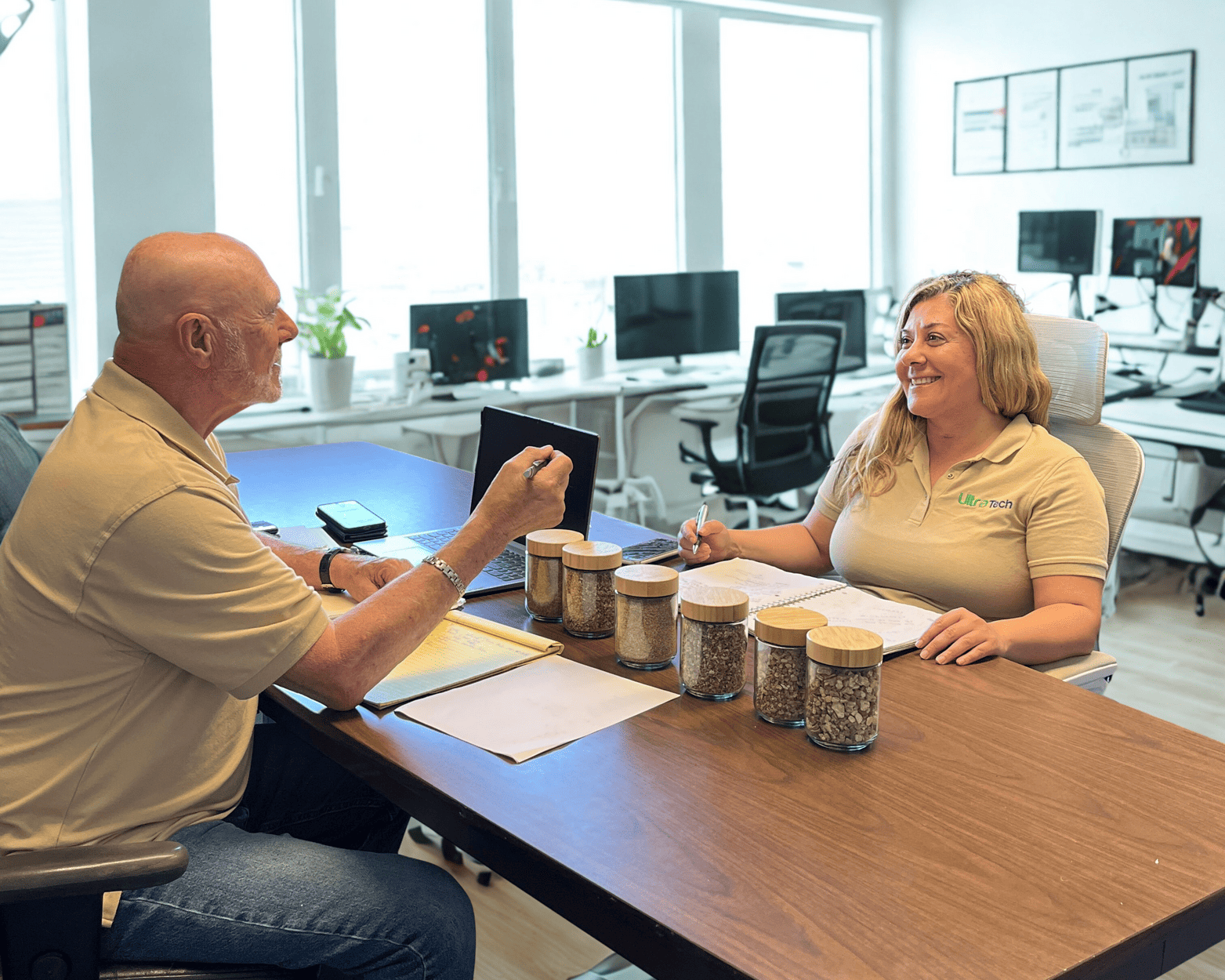 Two people in an office discuss samples in jars. Woman smiles. Man gestures. Modern office setting.