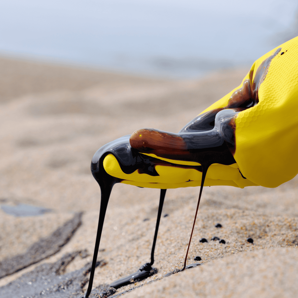 Yellow gloved hand covered in black oil on a sandy beach.