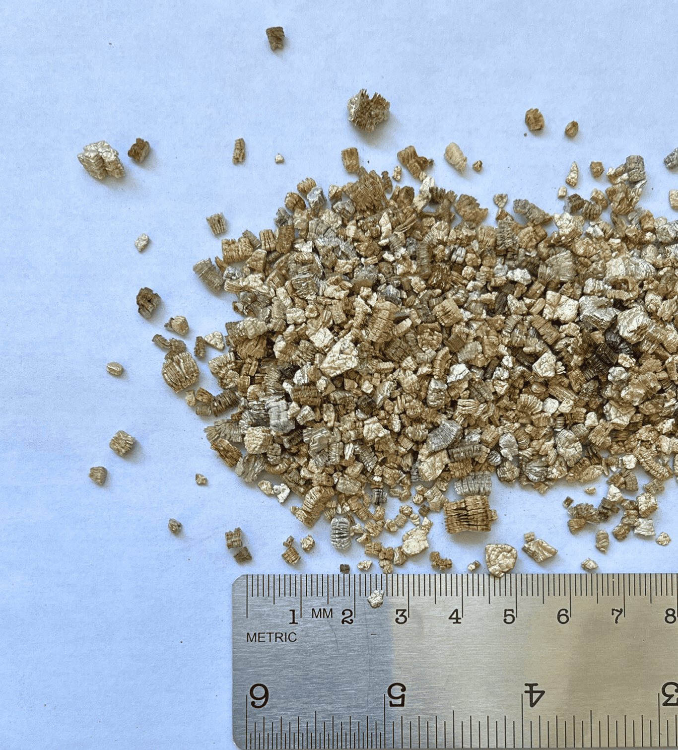 Pile of gold-colored vermiculite next to a ruler for scale, on a white surface.