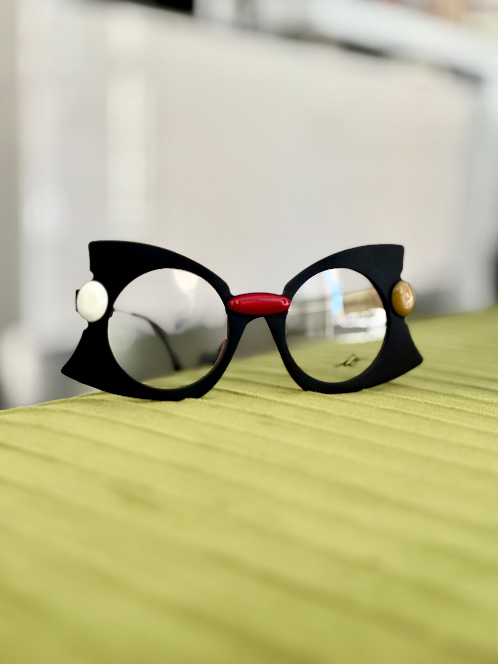 Occhiali neri con lenti circolari, ponte rosso e dettagli decorativi laterali, appoggiati su una superficie verde testurizzata.