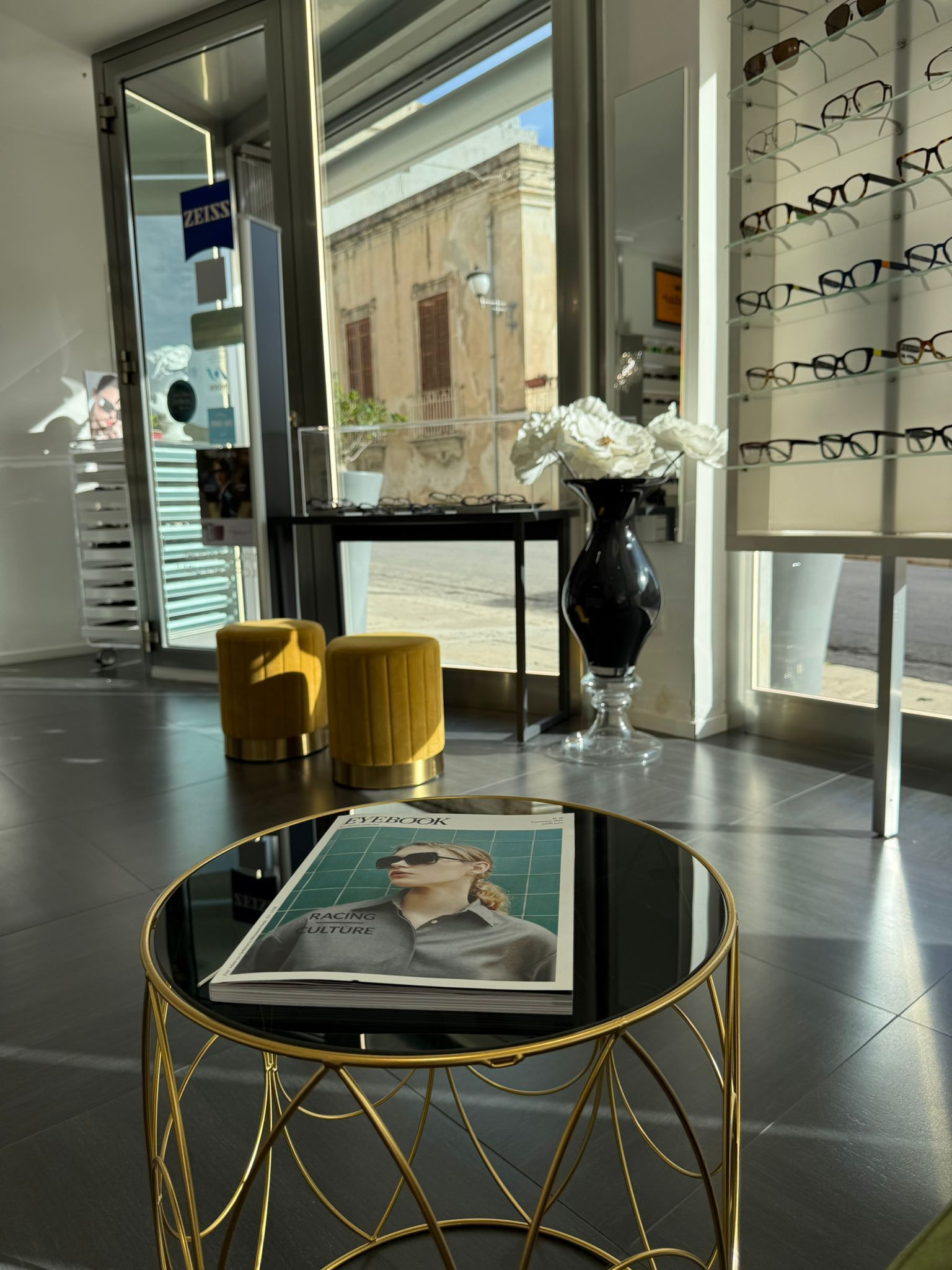 Interno di un negozio di ottica con tavoli espositivi, riviste e fiori. Arredamento giallo e oro. La luce del sole filtra attraverso la vetrina.