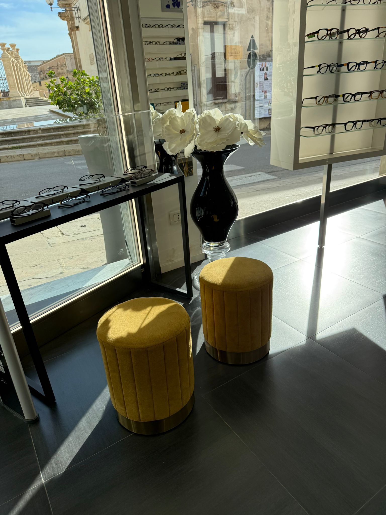 Sgabelli gialli, vaso nero con fiori bianchi e occhiali esposti vicino a una finestra soleggiata.