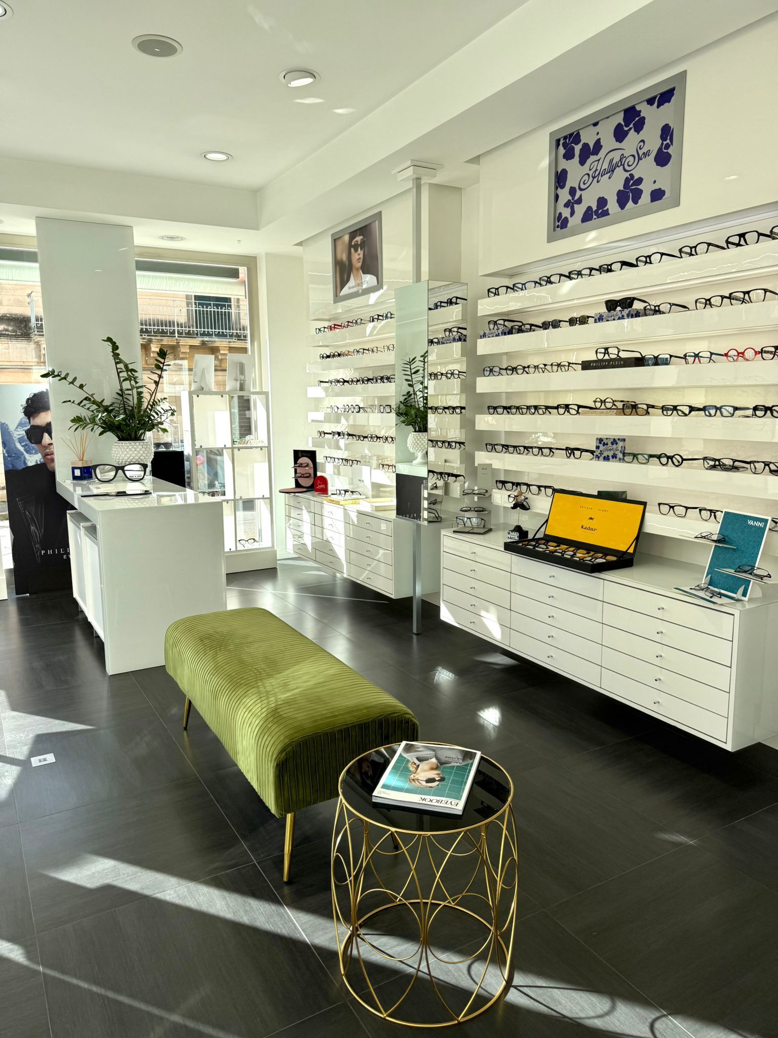 Interno di un negozio di occhiali. Pareti bianche, pavimento nero e scaffali con occhiali da vista. Panca verde e tavolino dorato.