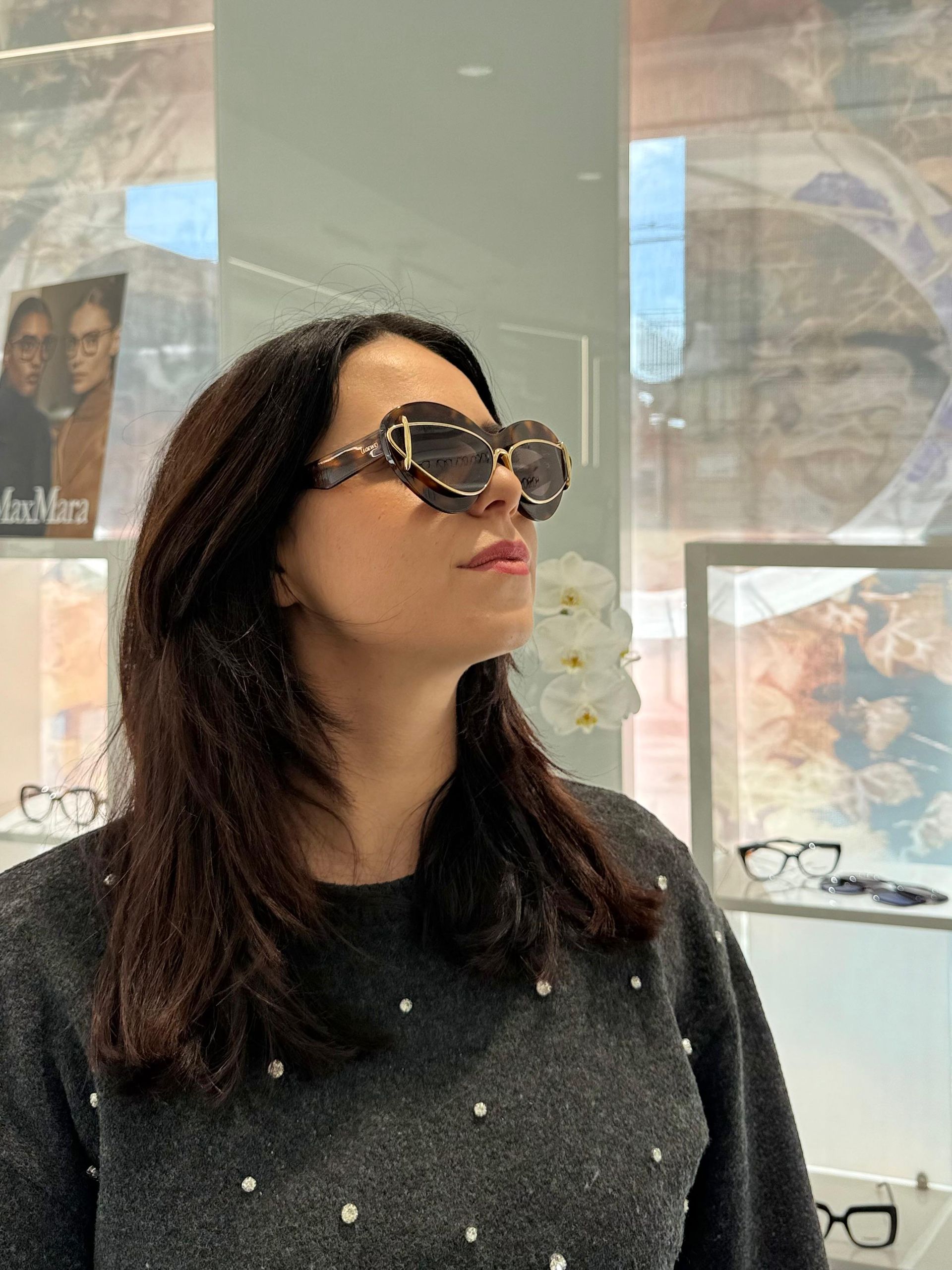 Donna che indossa grandi occhiali da sole fantasia in un ambiente interno, guarda verso l'alto; indossa un maglione grigio.