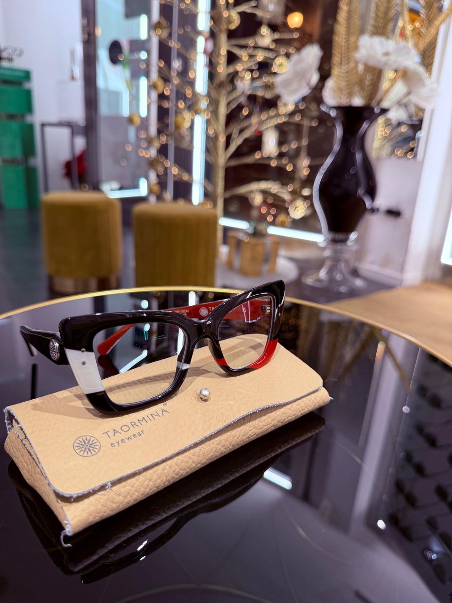 Occhiali con montatura nera e dettagli rossi su una custodia beige, appoggiati su un tavolo riflettente in un negozio di ottica.