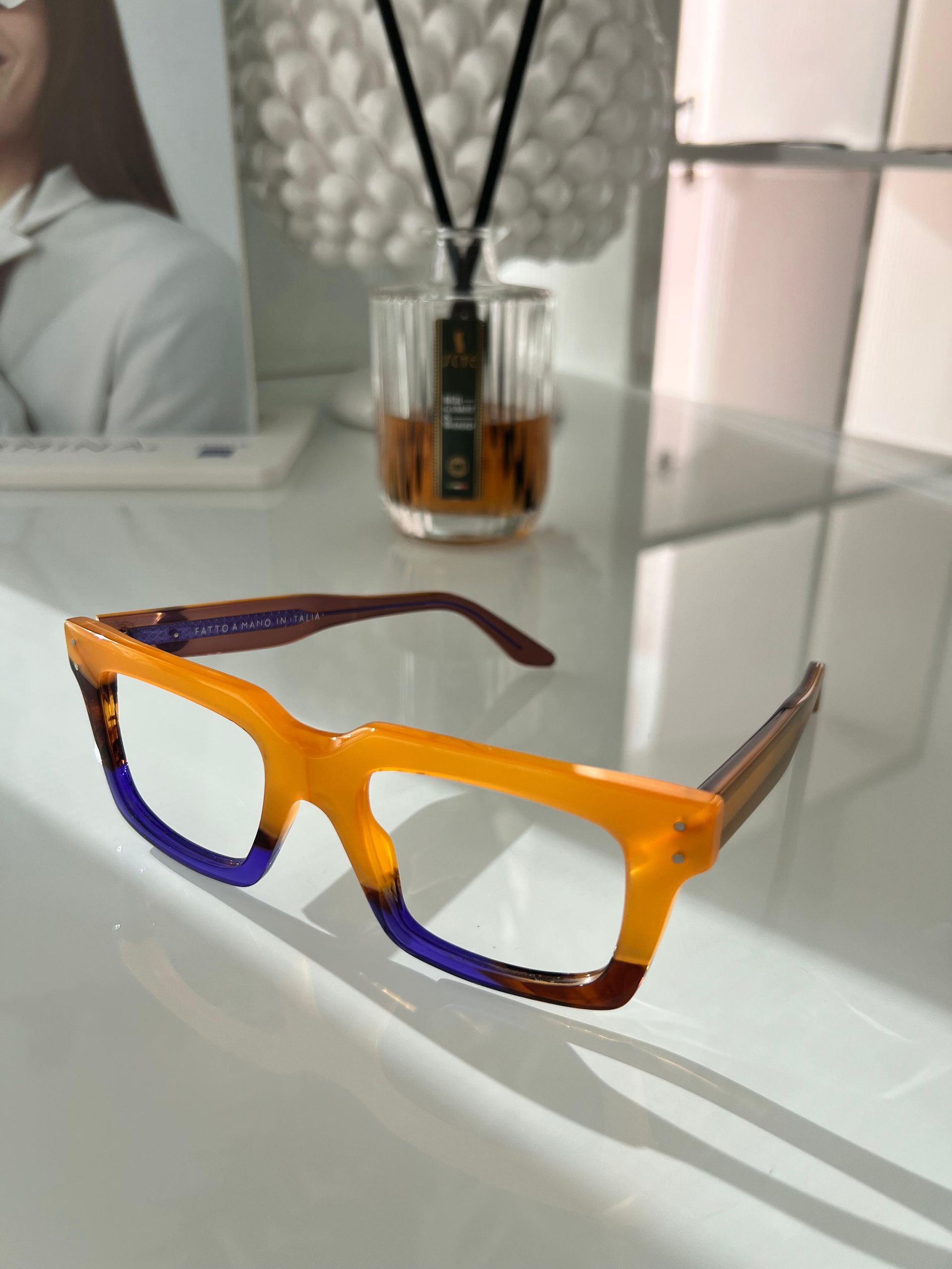 Occhiali arancioni e viola su una superficie bianca, con un bicchiere di liquido e un diffusore decorativo sullo sfondo.