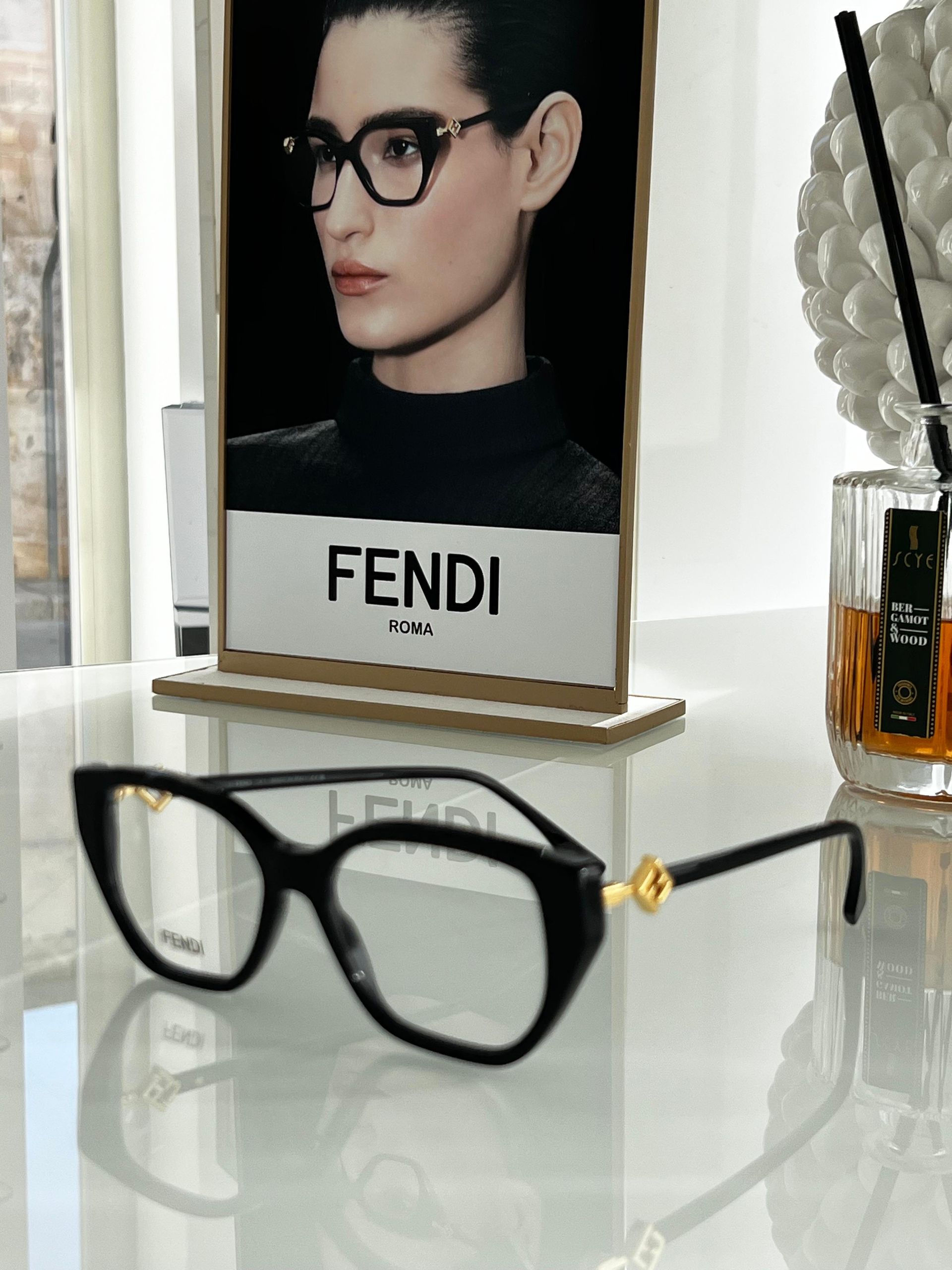 Occhiali da vista Fendi neri su un tavolo riflettente, con una pubblicità Fendi e un diffusore decorativo sullo sfondo.
