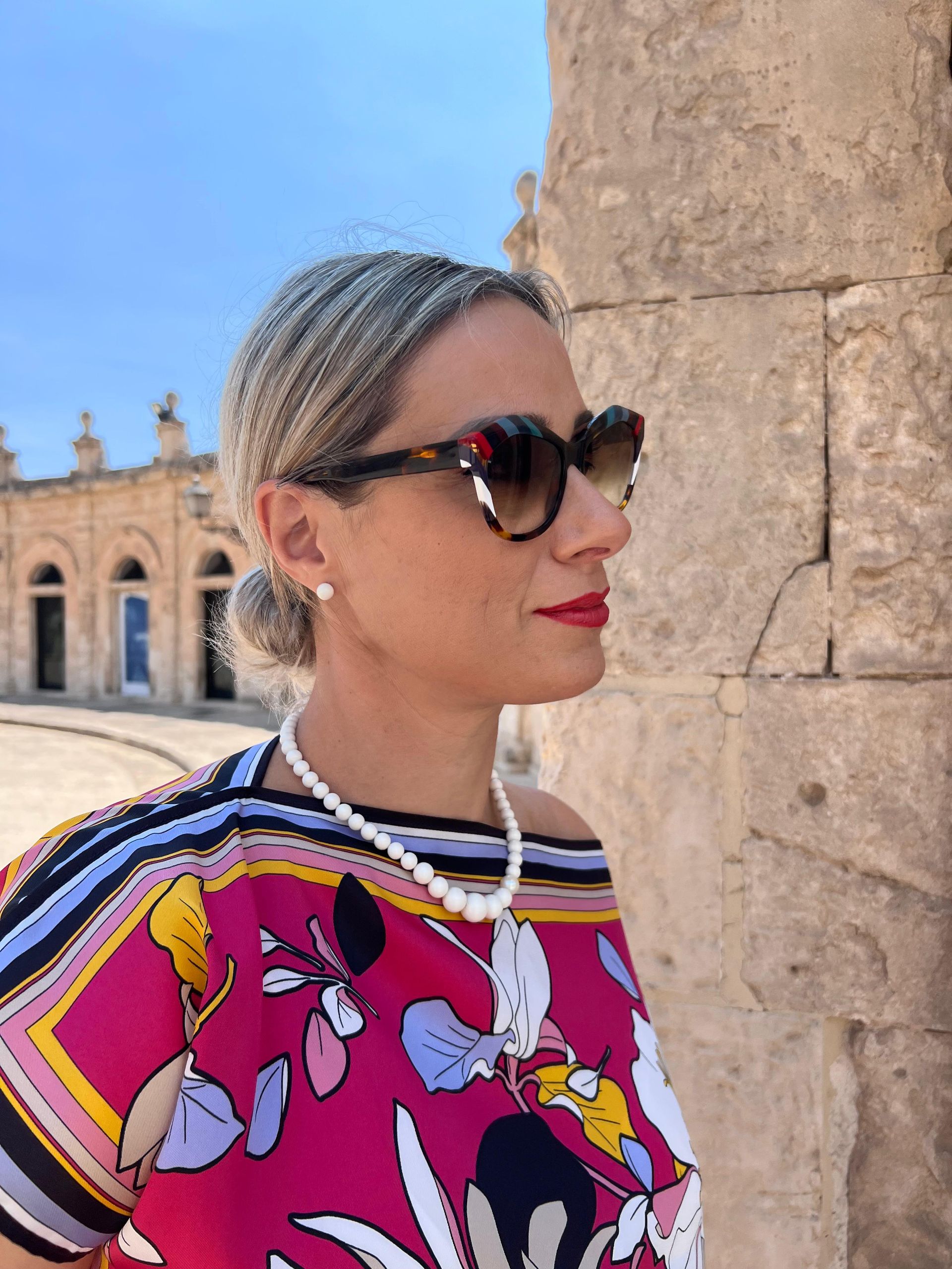 Una donna con occhiali da sole e un top colorato è in piedi vicino a una colonna di pietra all'aperto.