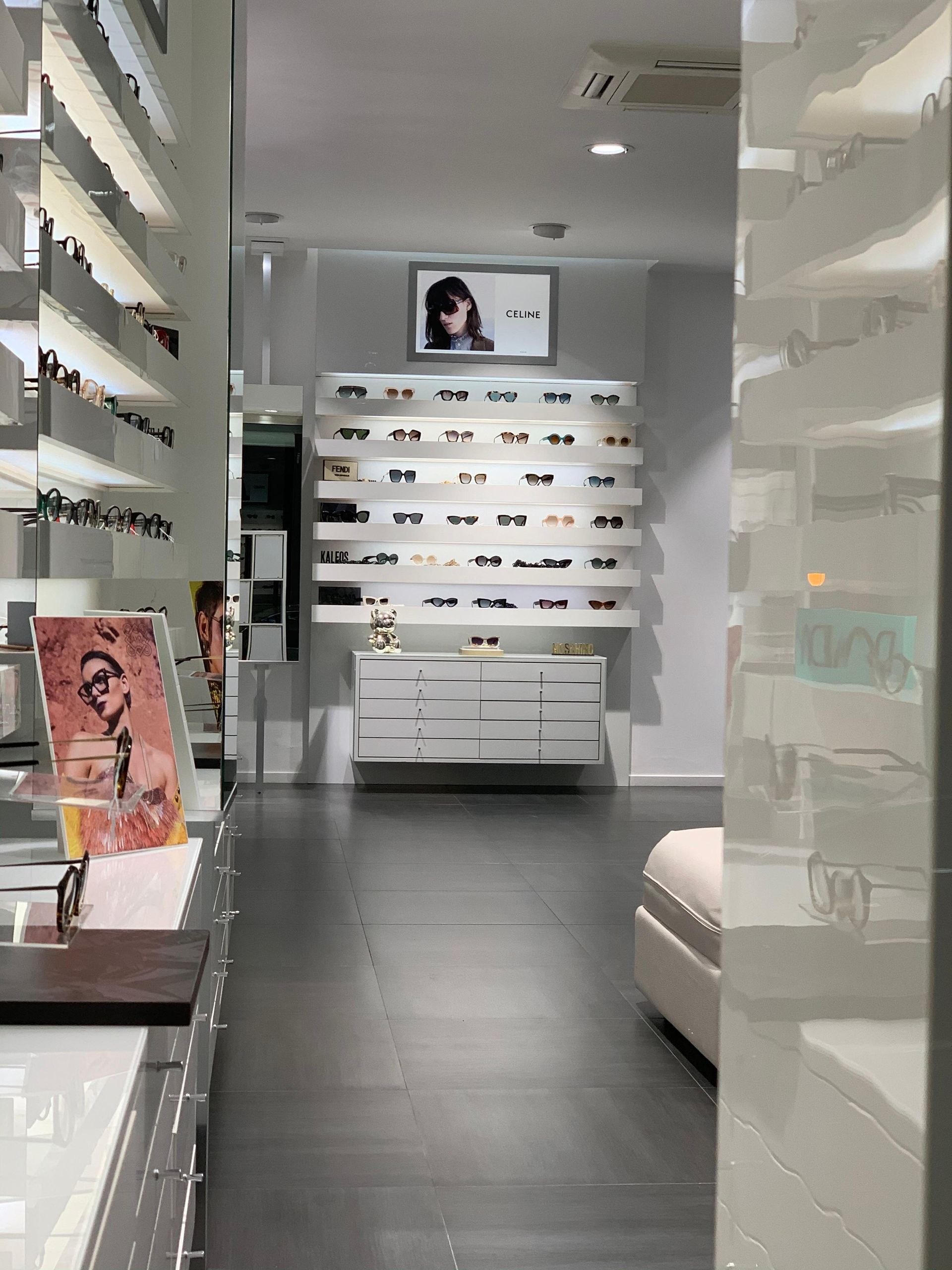 Interno di un negozio di occhiali; esposizione di occhiali da sole su scaffali e vetrine bianche; pavimento nero.