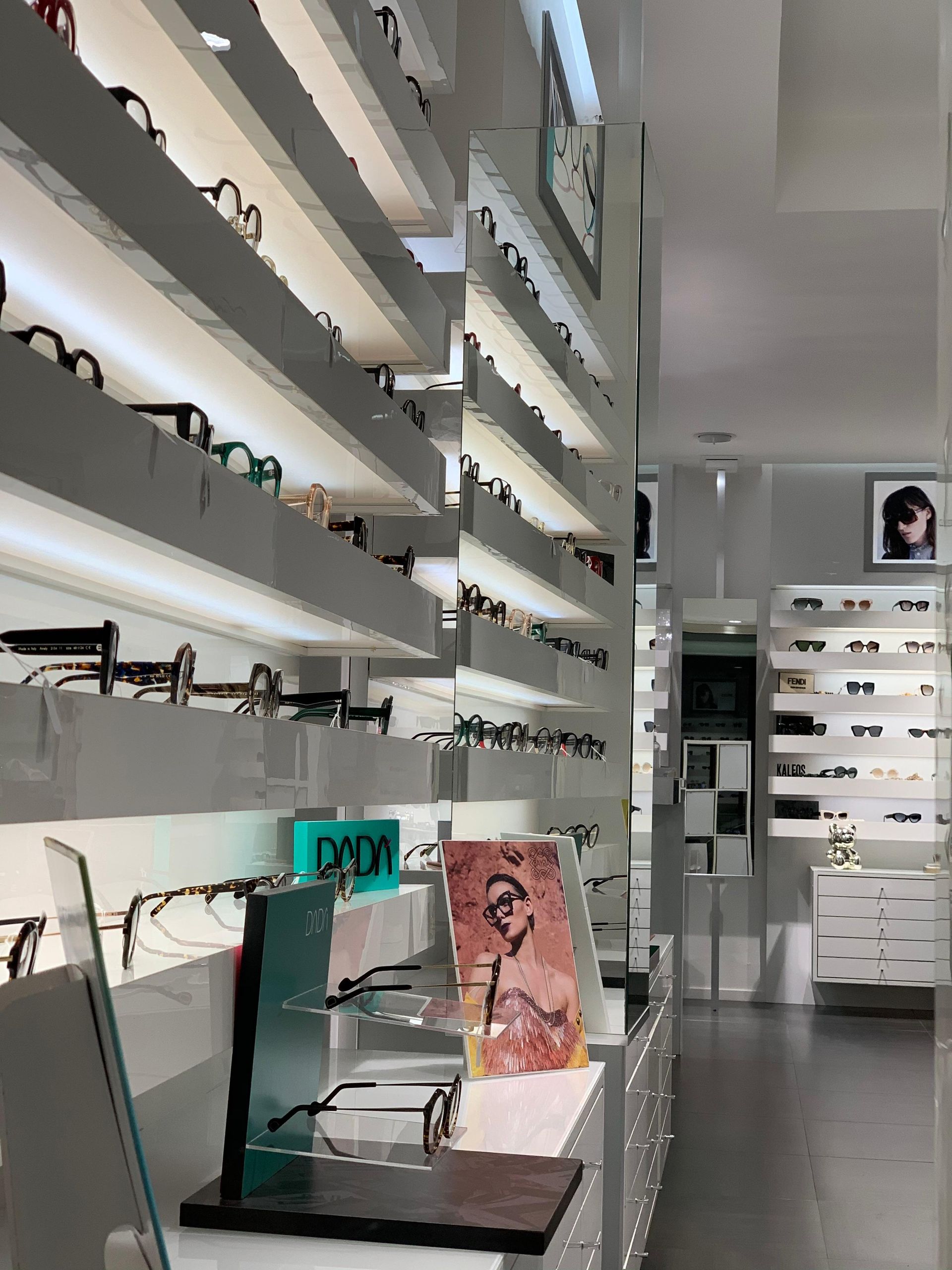 Interno di un negozio di occhiali; scaffali con numerosi occhiali, luci intense, decorazioni bianche e grigie.
