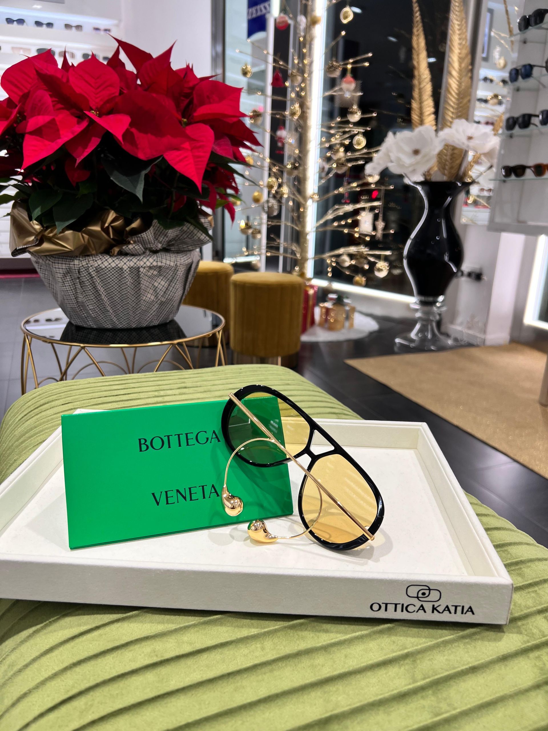 Occhiali da sole su un vassoio bianco con un cartoncino verde, vicino a stelle di Natale rosse in un negozio.