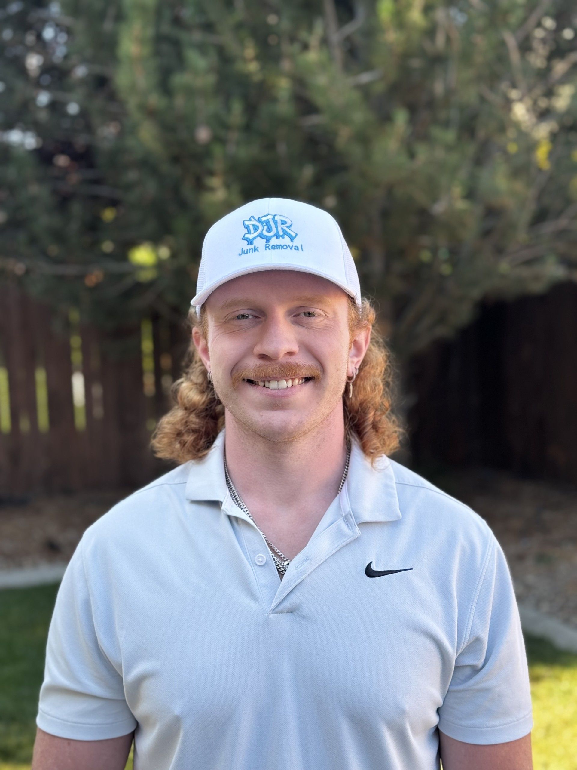 Man with red hair and mustache wearing a white hat and gray polo shirt smiling outdoors.