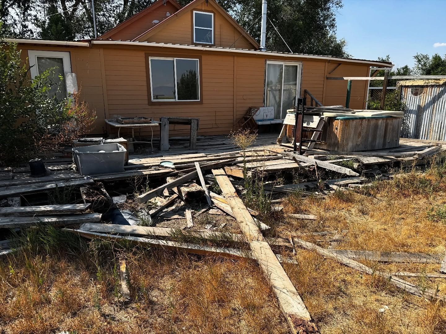 Dilapidated brown house with a destroyed wooden deck and overgrown weeds.