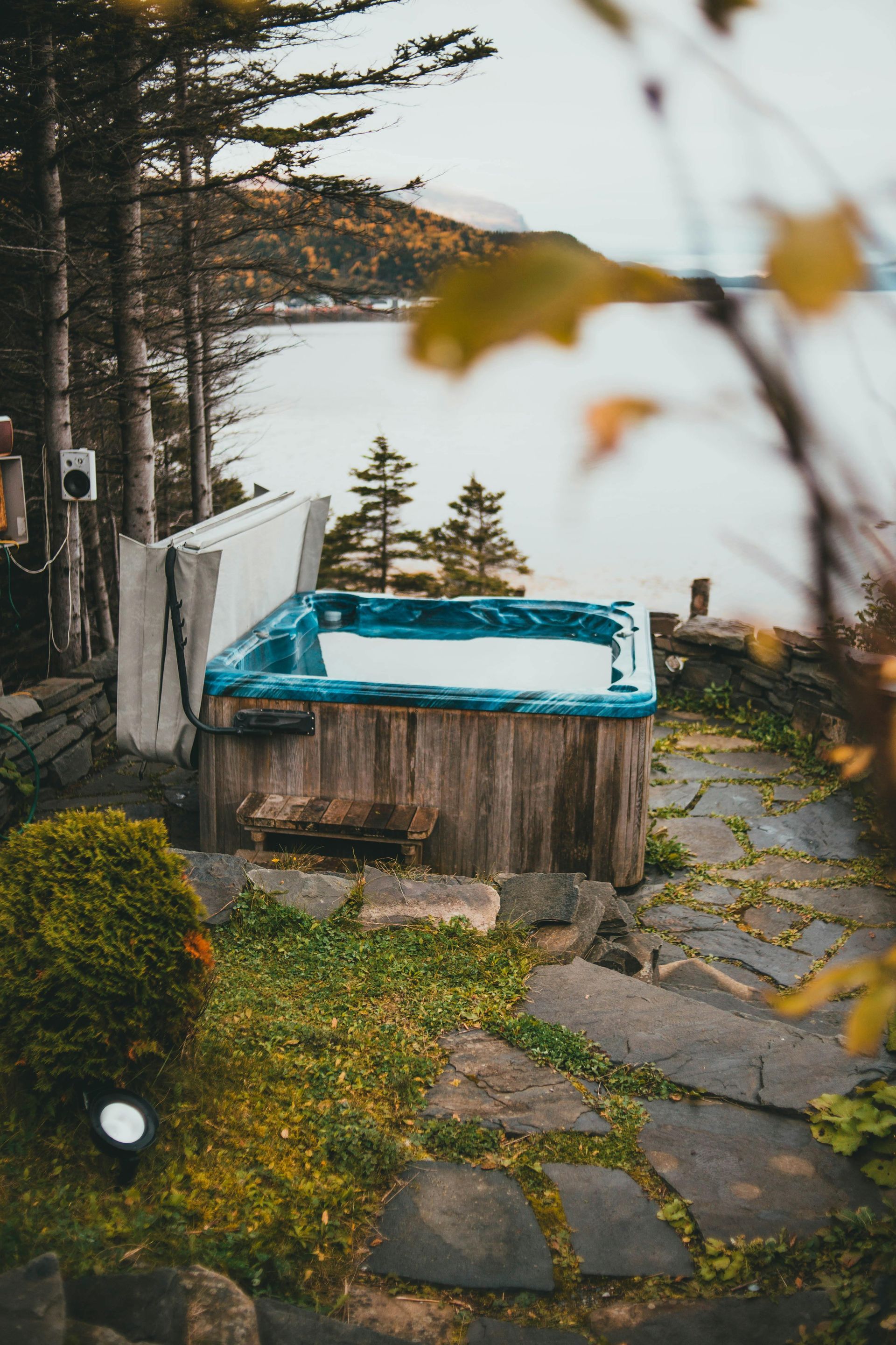 Wooden hot tub with blue interior sits outdoors near a body of water, foliage in foreground and trees in background.