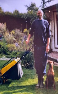 A person standing on a lawn with a yellow lawnmower and a brown dog next to a garden and a house.