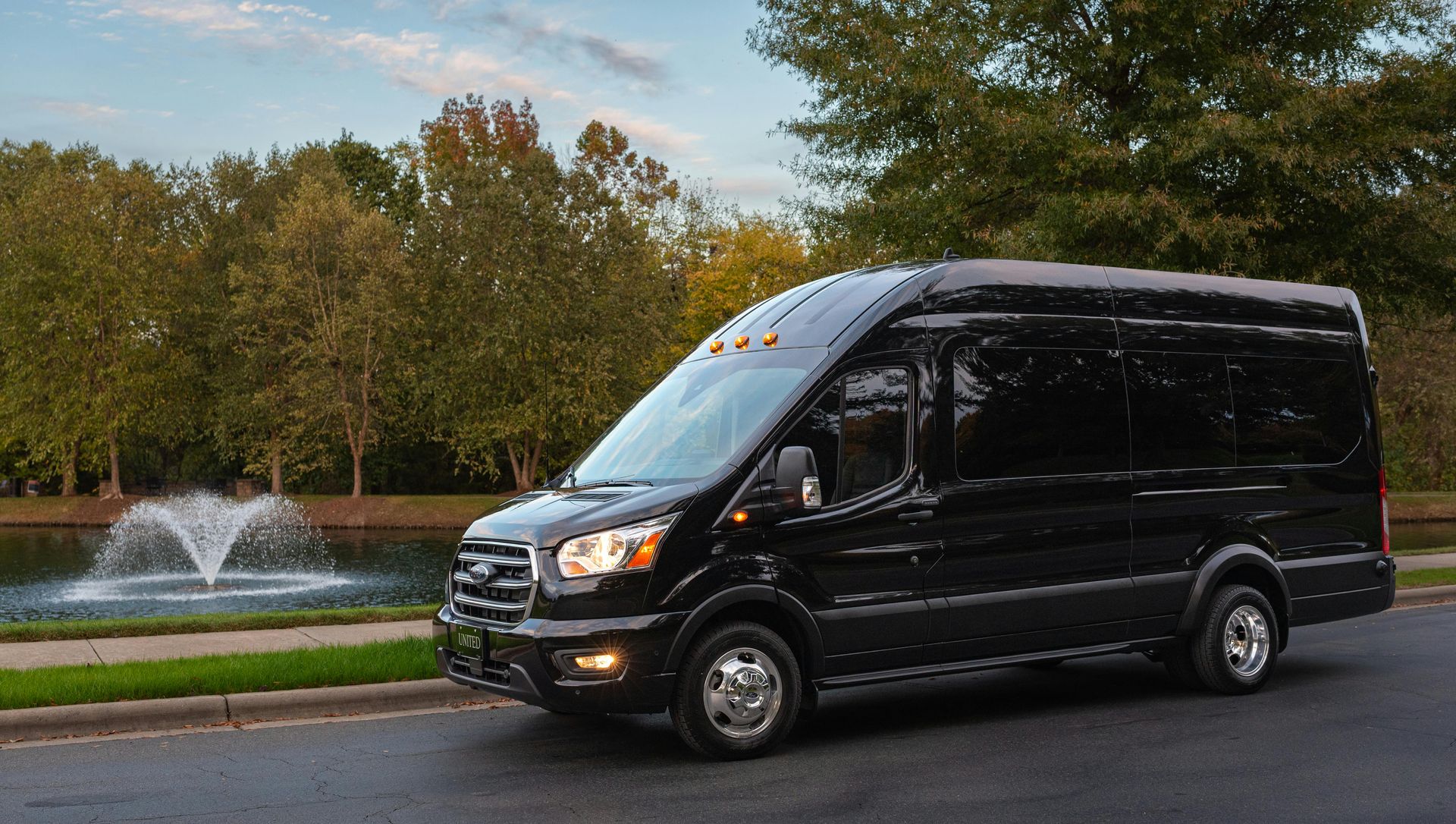 A black Ford Transit passenger van parked on a road next to a park with a fountain in the background.