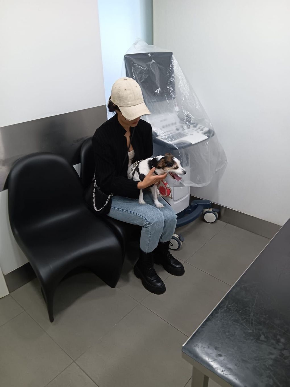 A person in a baseball cap and dark jacket sits on a black chair, holding a small dog in a clinical, white-walled room.