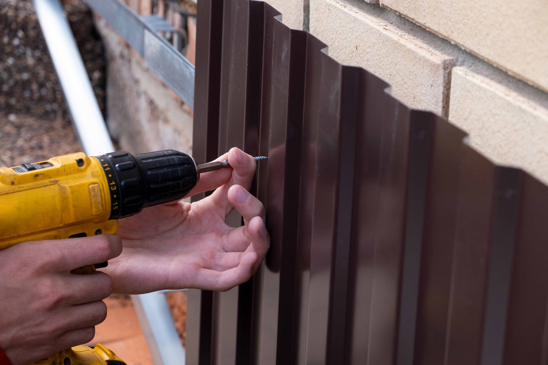Person using a yellow drill to screw a panel into a brick wall; brown metal panel and brown screw are visible.