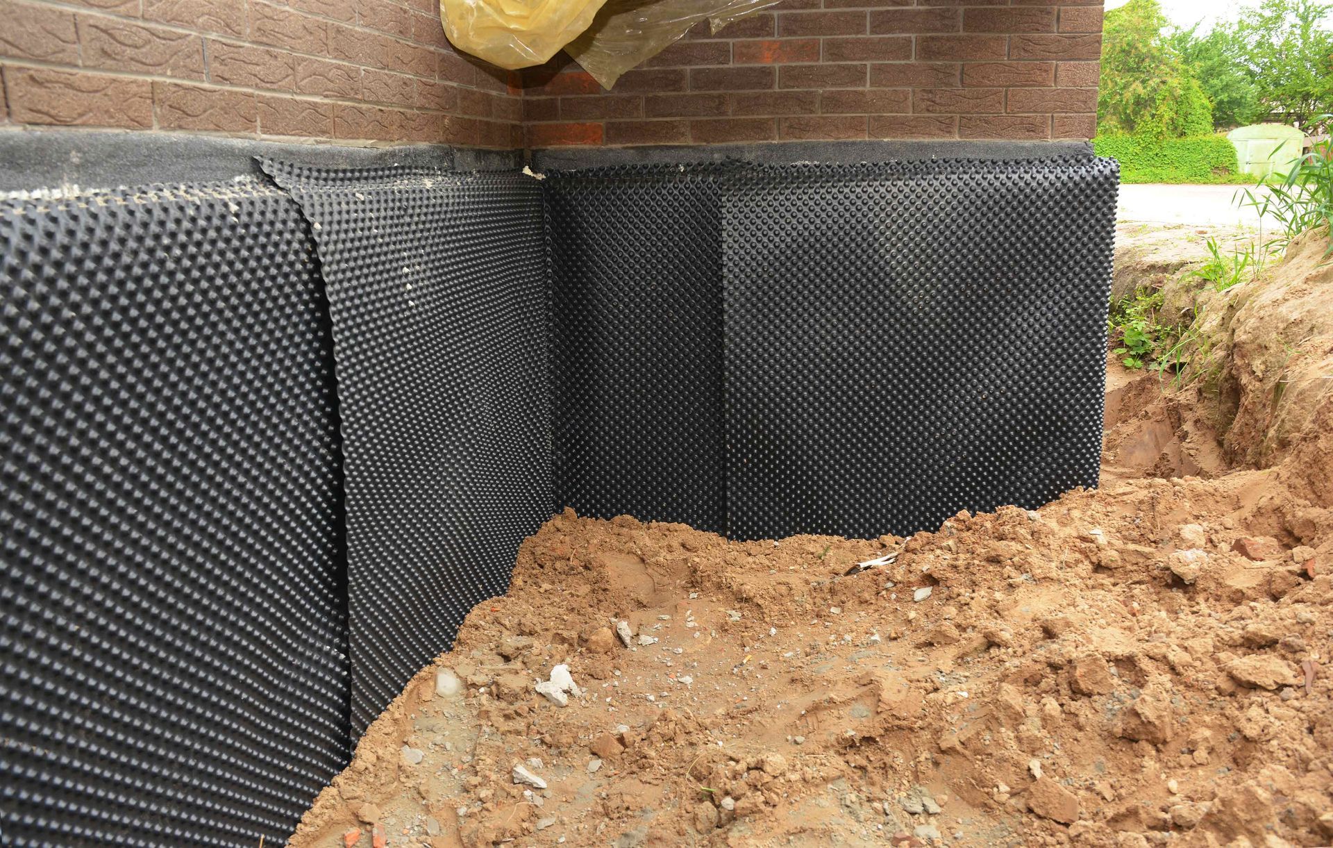 Black dimpled drainage membrane installed on a brick foundation wall with surrounding soil.