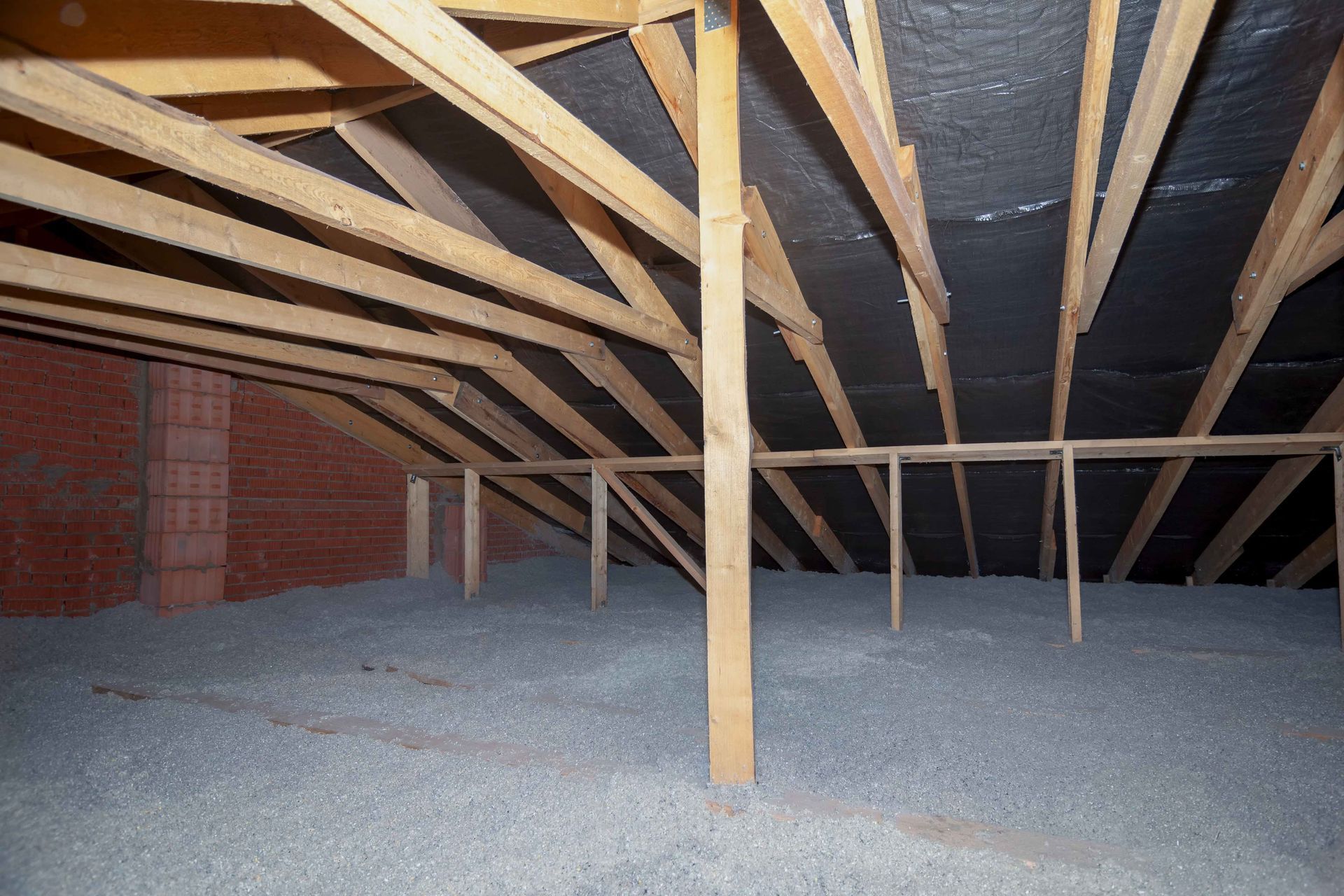 Attic with wooden beams, brick wall, and insulation on the floor. Black material is secured to the rafters.