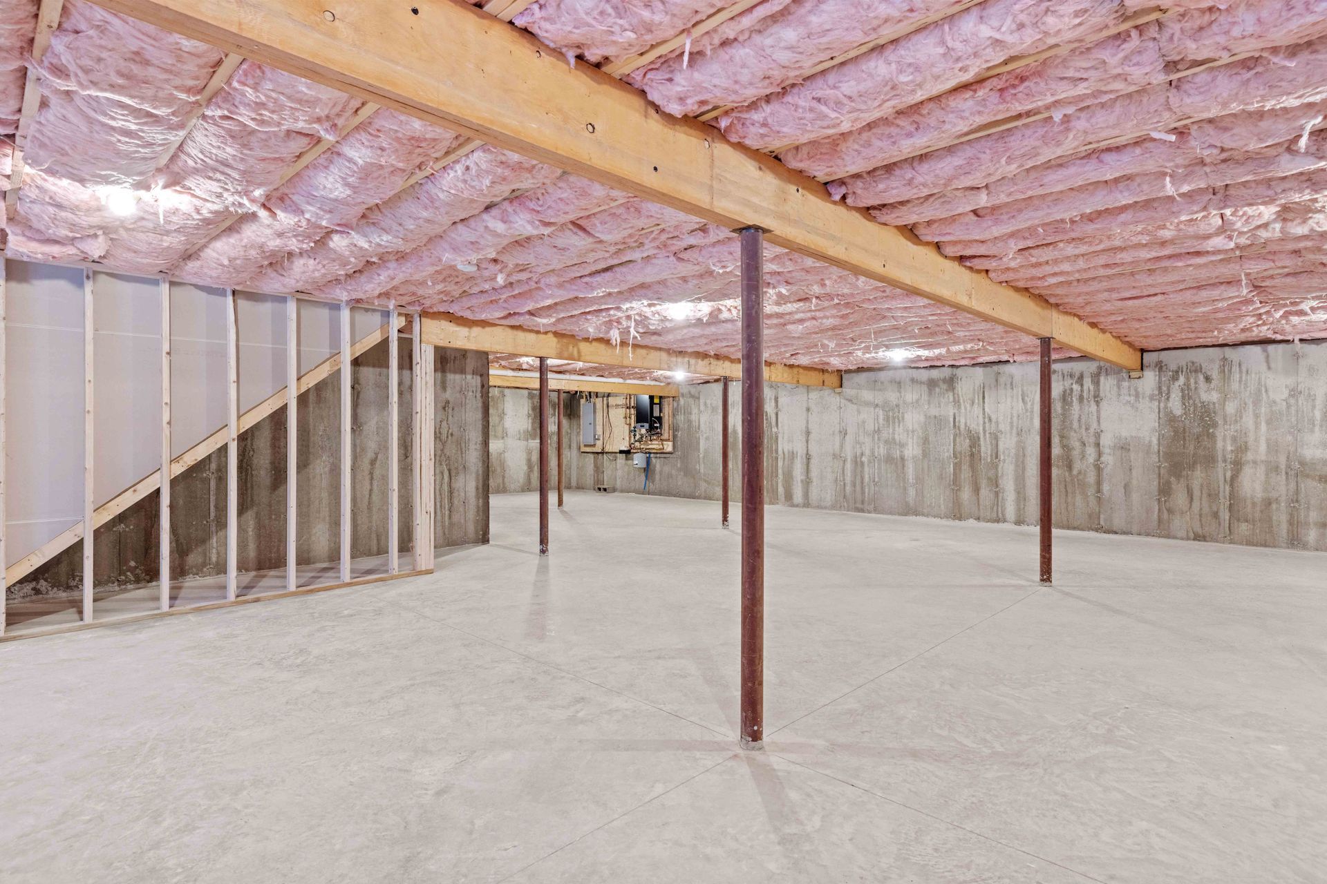 Unfinished basement with exposed ceiling insulation, wooden beams, concrete walls and floor, and support beams.