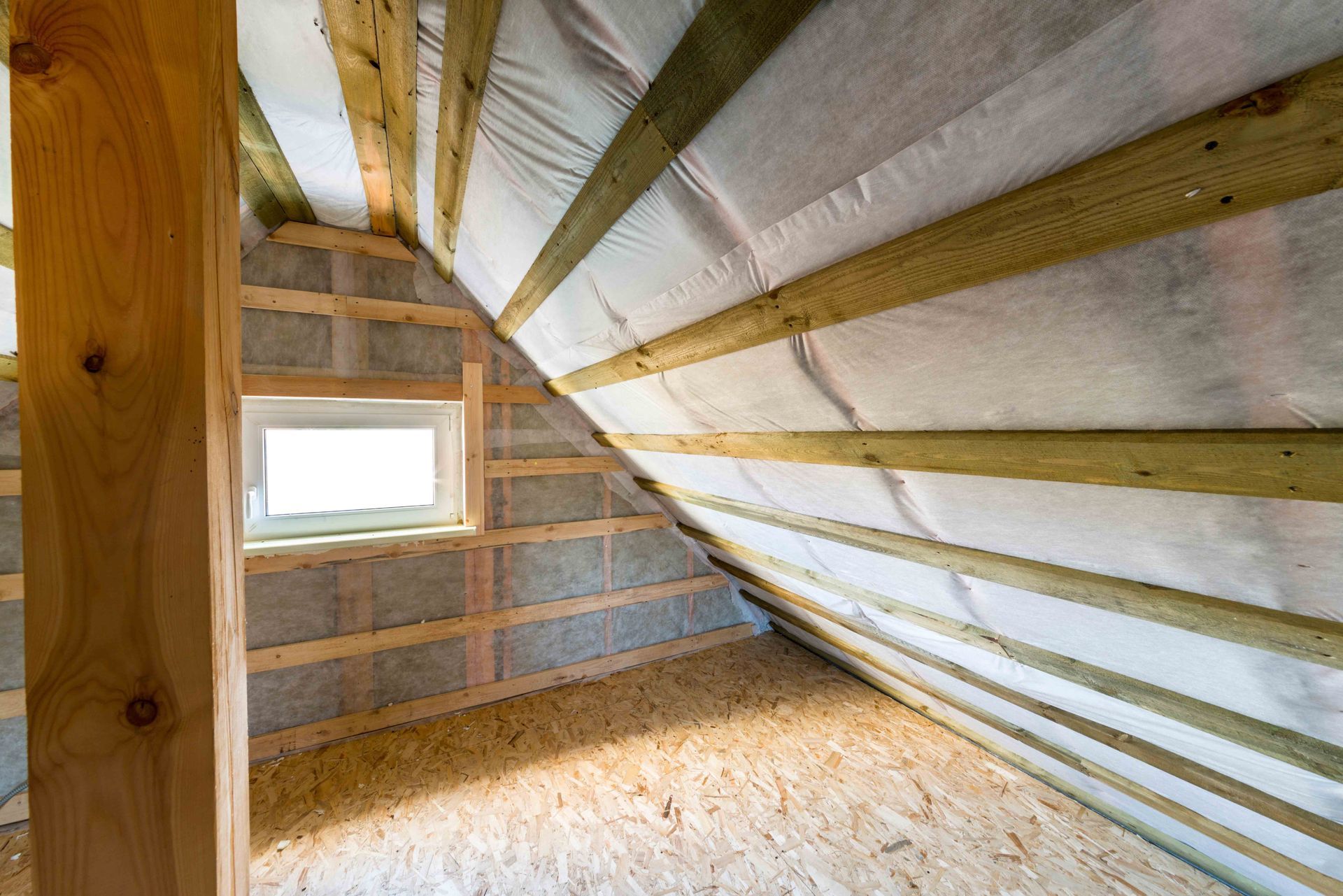 Interior view of an attic space with exposed wood beams, insulation, cinder block walls, and a small window.
