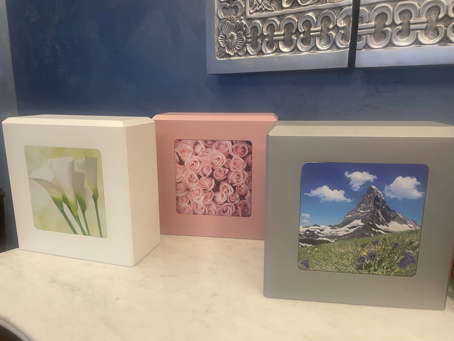 Tre scatole quadrate: bianca con calle, rosa con rose e grigia con un paesaggio montano.