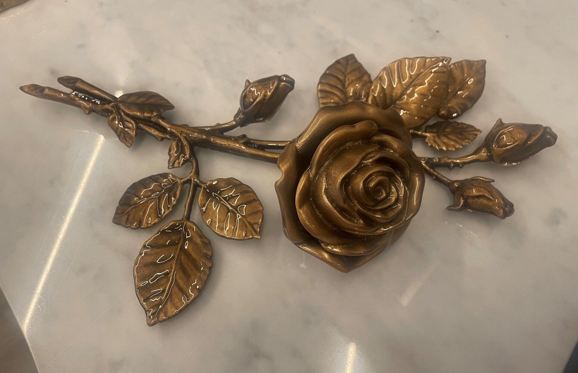 Scultura di rosa color oro con foglie e boccioli dettagliati su una superficie di marmo bianco.