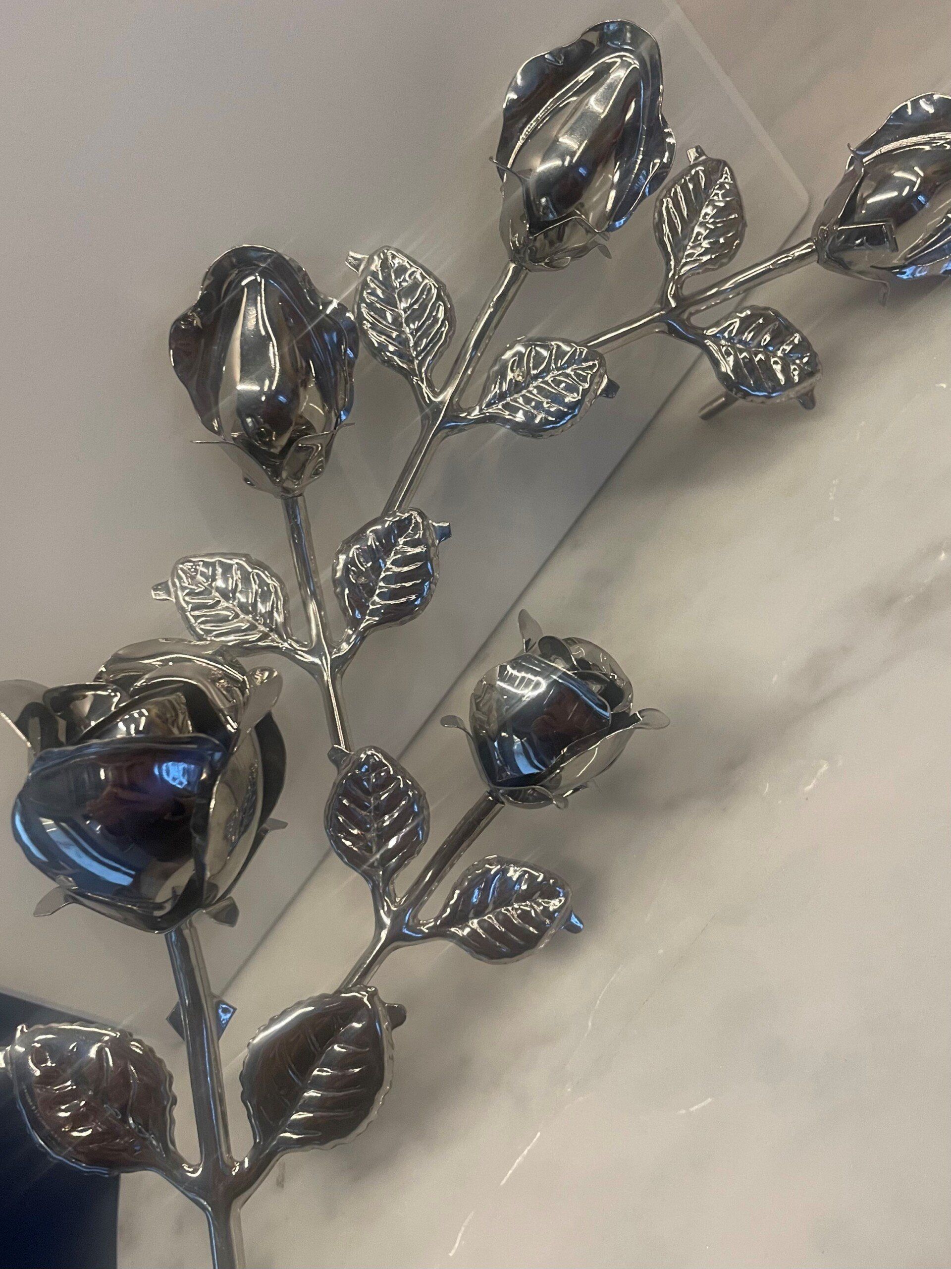 Stelo di rosa in metallo color argento con boccioli di fiori chiusi e foglie, appoggiato su una superficie bianca.