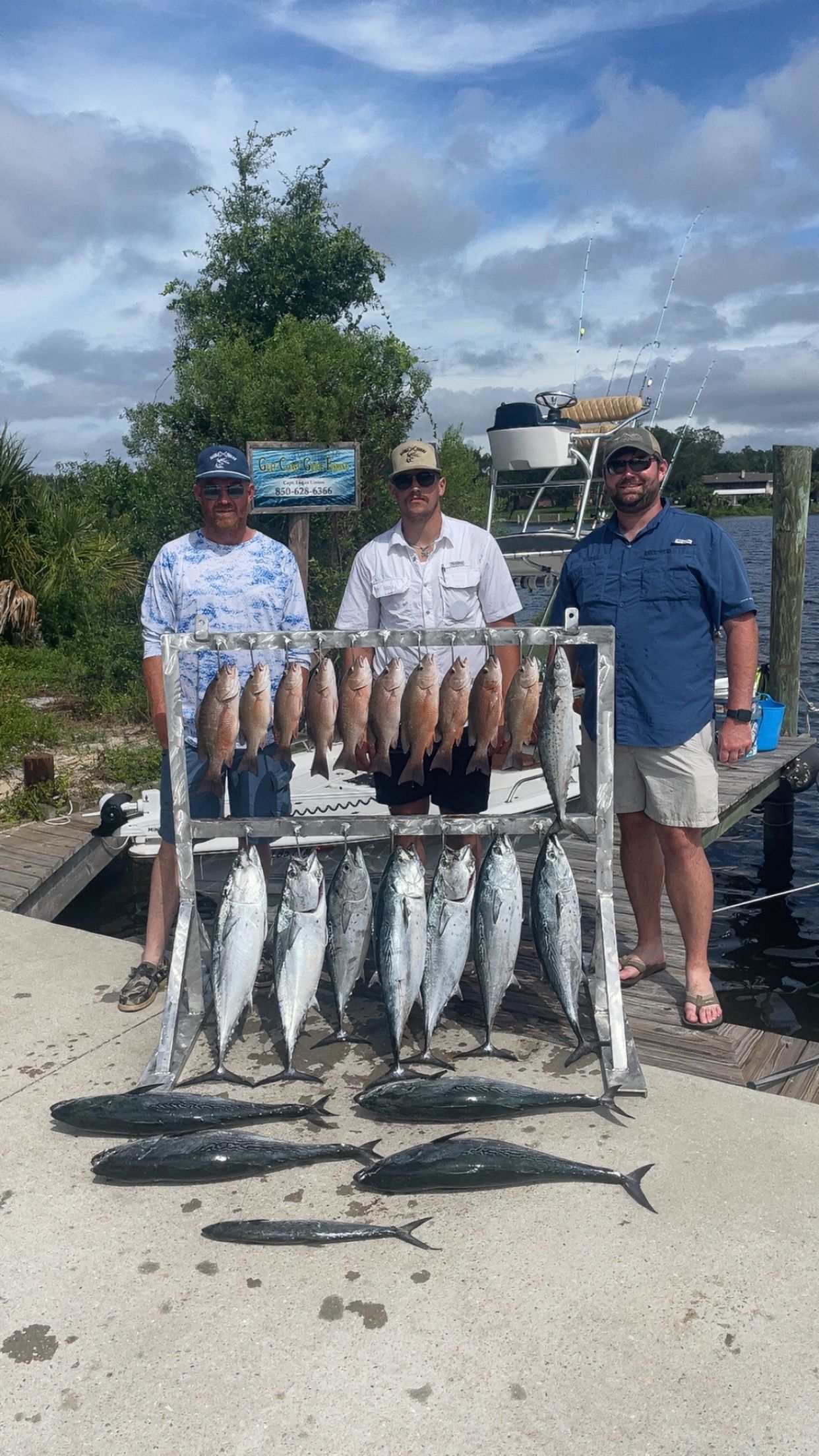 Three men pose with a metal rack displaying various fish at a dock, under a partly cloudy sky.